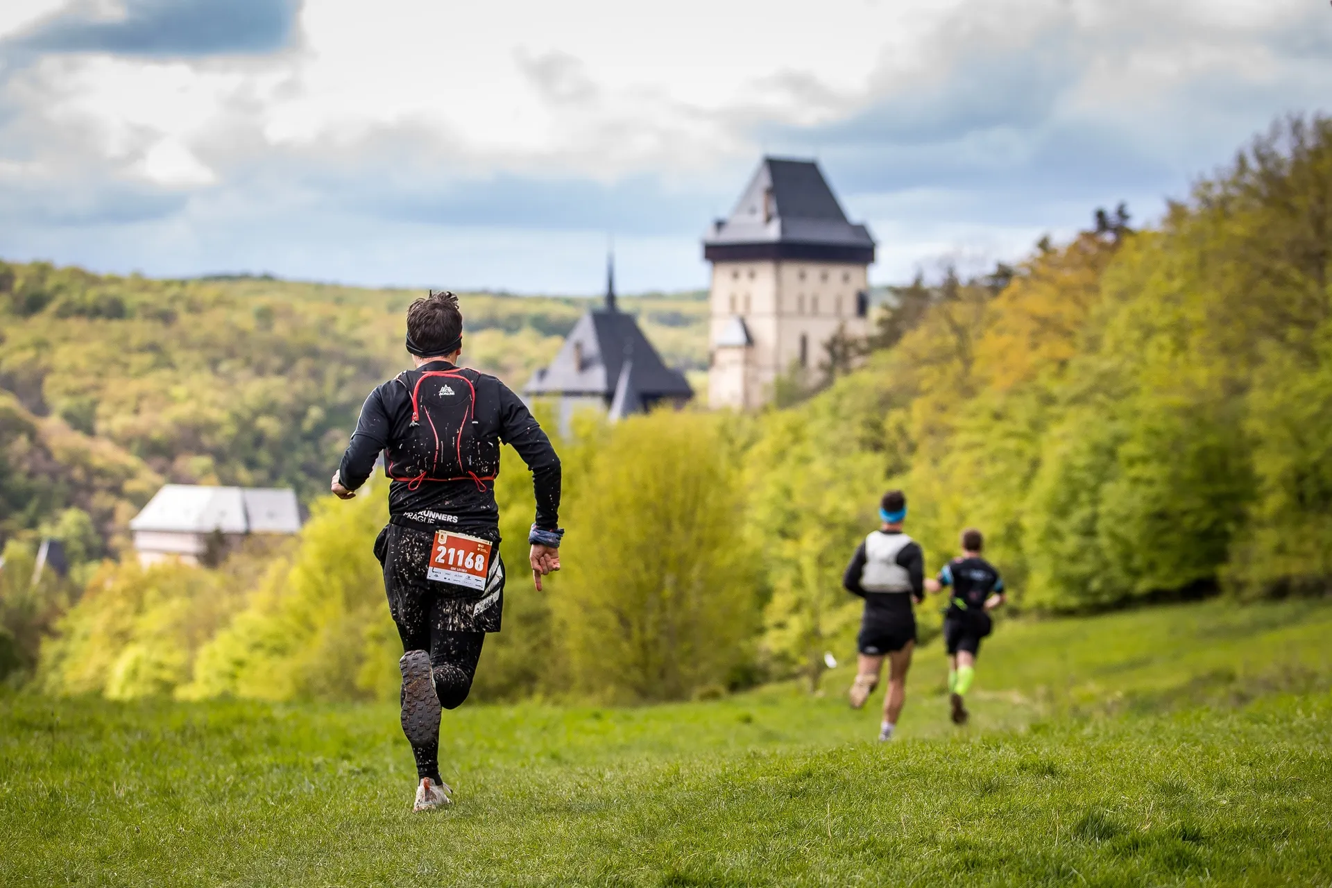 The image shows three people running on a grassy trail. One person is in the foreground, wearing a backpack and a race number on their back. In the background, there's a castle-like building surrounded by trees. The scene appears to take place in a scenic, forested area.
