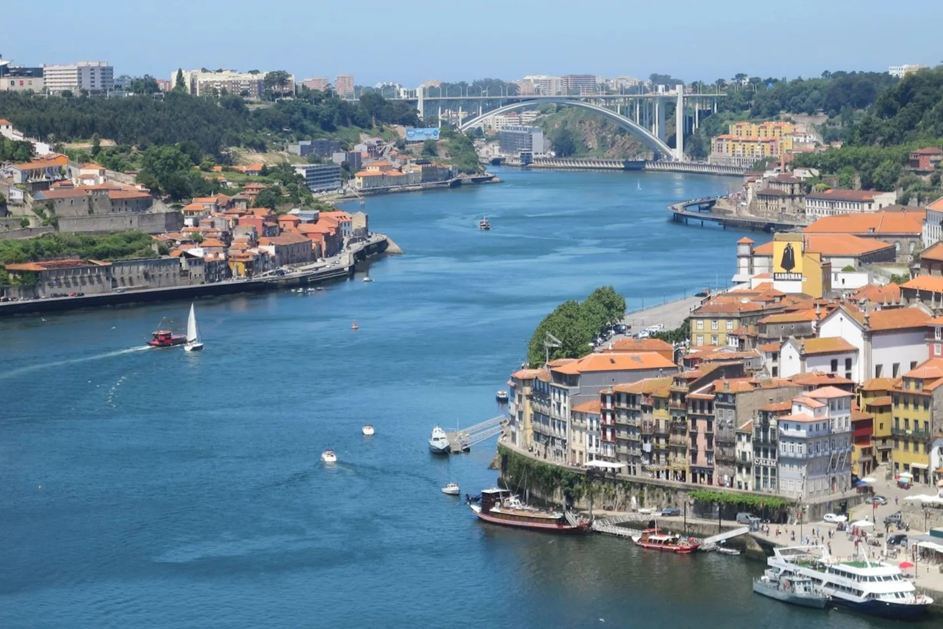 The image shows a panoramic view of a city with a river running through it. There are several buildings with varied architecture along the riverbanks, suggesting a dense urban area. A distinctive bridge with an arch design spans the river, connecting the two sides of the city. There are boats on the water, including one that seems to be a tour or passenger boat. The terrain on the far side of the river rises into what appears to be a hill or cliff with vegetation and buildings on it. This is indicative of a city with a significant history and development, likely in Europe due to the architectural style and bridge design.