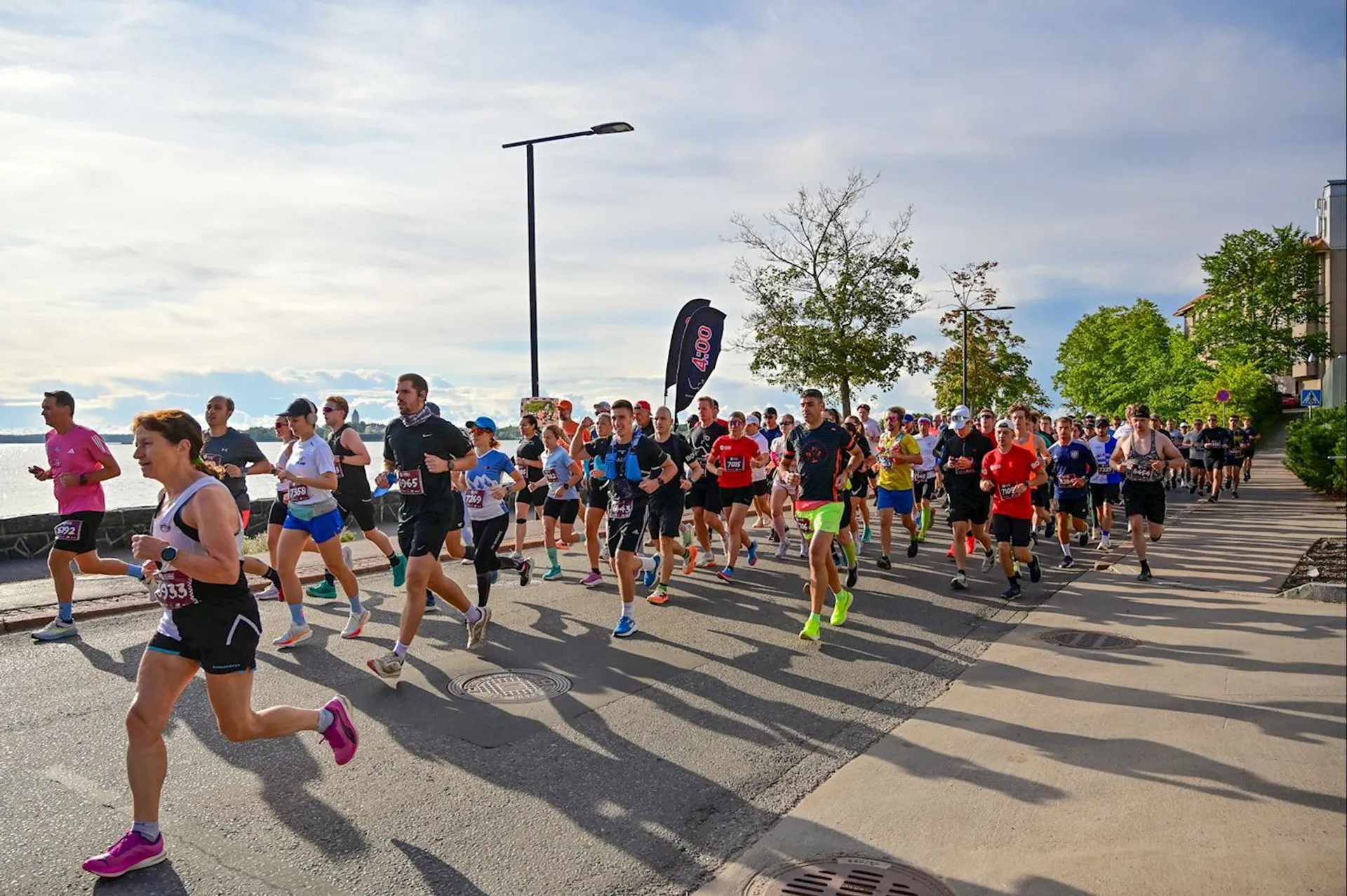The image shows a group of people participating in a road race. They are running along a street next to a body of water with trees and a few buildings in the background. The sky is clear and sunny.