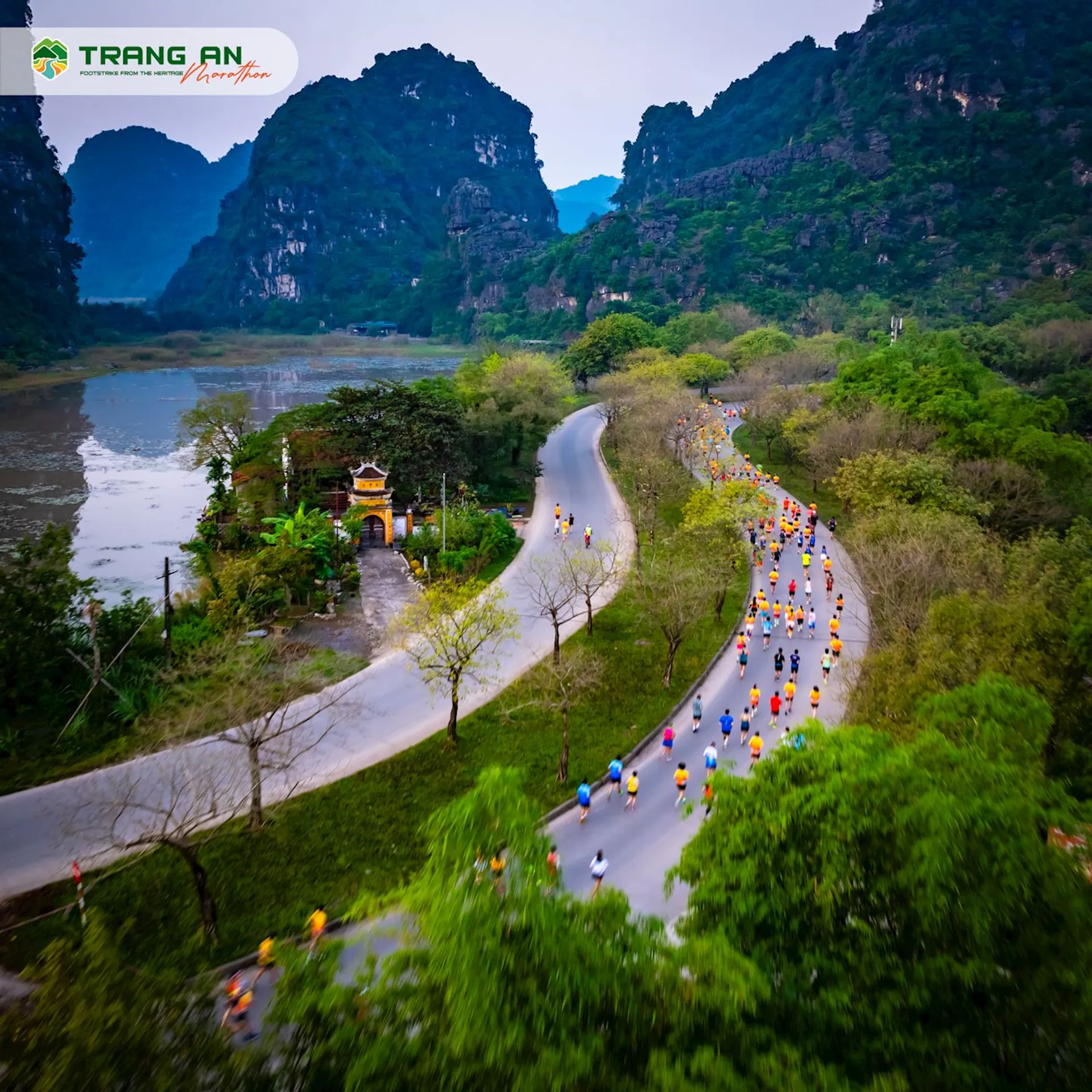 The image shows a scenic view with a winding road running through a lush, green landscape dotted with limestone karst mountains. There is a considerable number of people on the road, appearing to be cyclists and pedestrians possibly participating in an event or just enjoying the outdoor activities in a picturesque setting. A small structure, which could be a pagoda or shrine, is visible at the road's edge near the river. The area has a tranquil vibe, with the waterbody on one side of the road likely adding to the serene atmosphere of the place. The text "Trang An" suggests that this could be the Trang An Landscape Complex, a UNESCO World Heritage site in Vietnam, known for its stunning natural beauty and cultural significance.