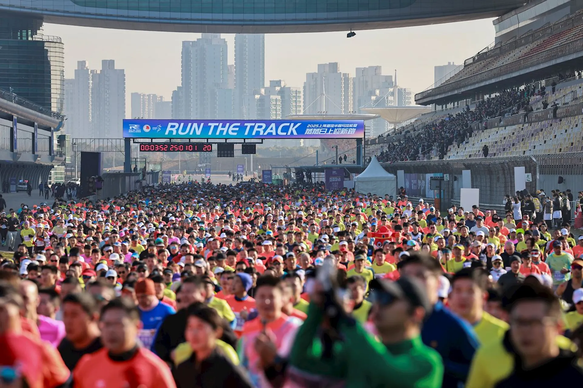 The image shows a large group of people participating in a running event on a track. There is a banner overhead that says "RUN THE TRACK." The background depicts a cityscape with tall buildings. The participants are wearing colorful athletic attire.