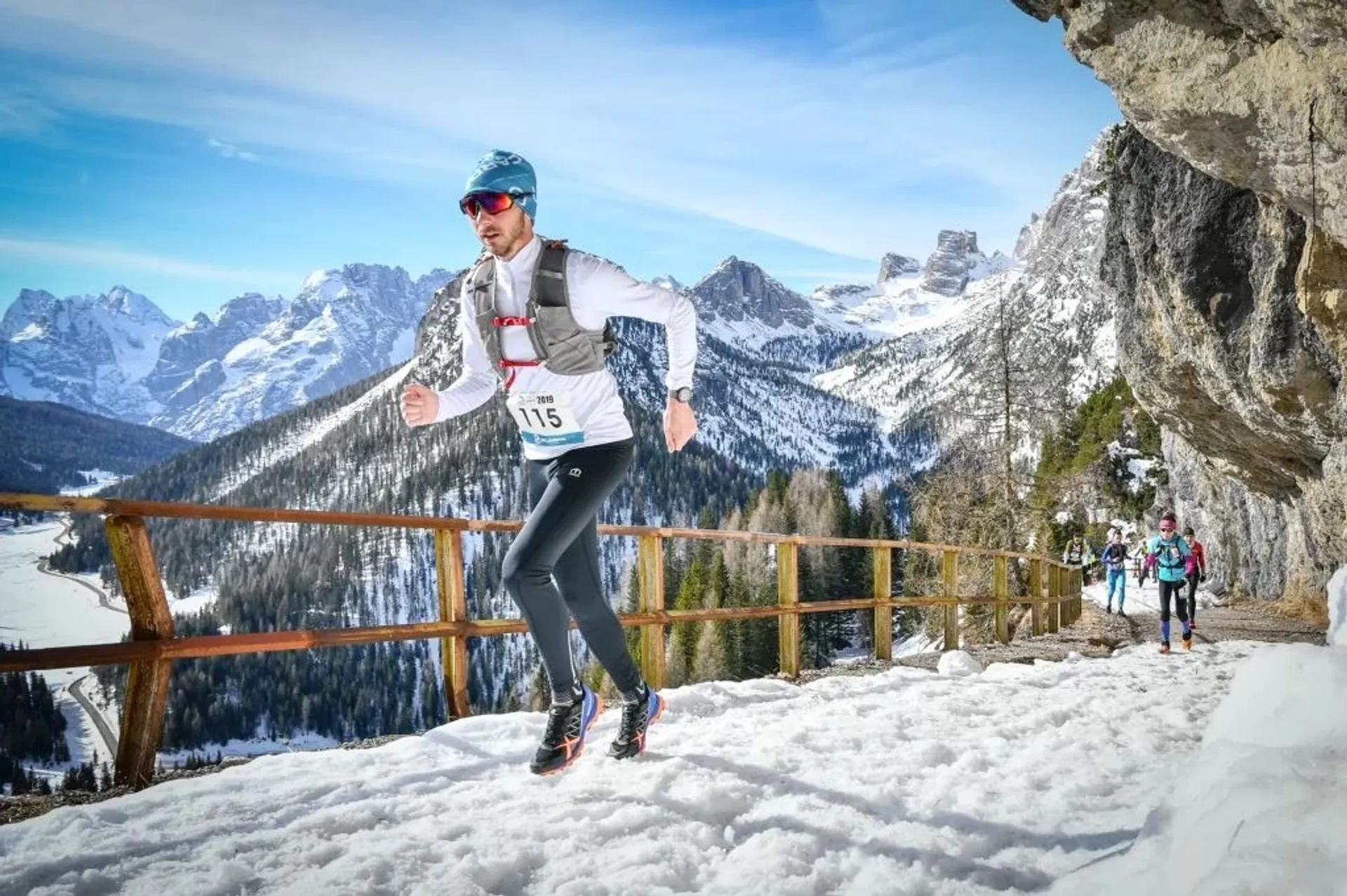 This image shows a trail running competition, likely a winter or mountain race, given the snowy conditions. The focus is on a male runner wearing a race bib labeled "115," who is running vigorously on a snow-covered trail. He is dressed in running tights, a long-sleeved shirt, a beanie, and sunglasses, and he has a hydration pack on his back. The runner has a watch on his left wrist, possibly a GPS watch to track his performance.

Behind him, there's at least one other competitor following the same trail. The background reveals a stunning mountain landscape with snow-capped peaks under a clear blue sky, suggesting that the race is set in a high alpine environment.

The participants are equipped appropriately for cold weather