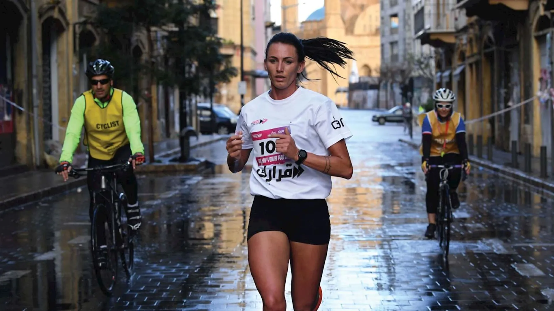 The image shows a woman running in a race on a wet street, wearing a bib with the number 10. She is accompanied by two cyclists on either side. The setting appears to be an urban area with buildings visible in the background. The ground is reflective, indicating recent rain.