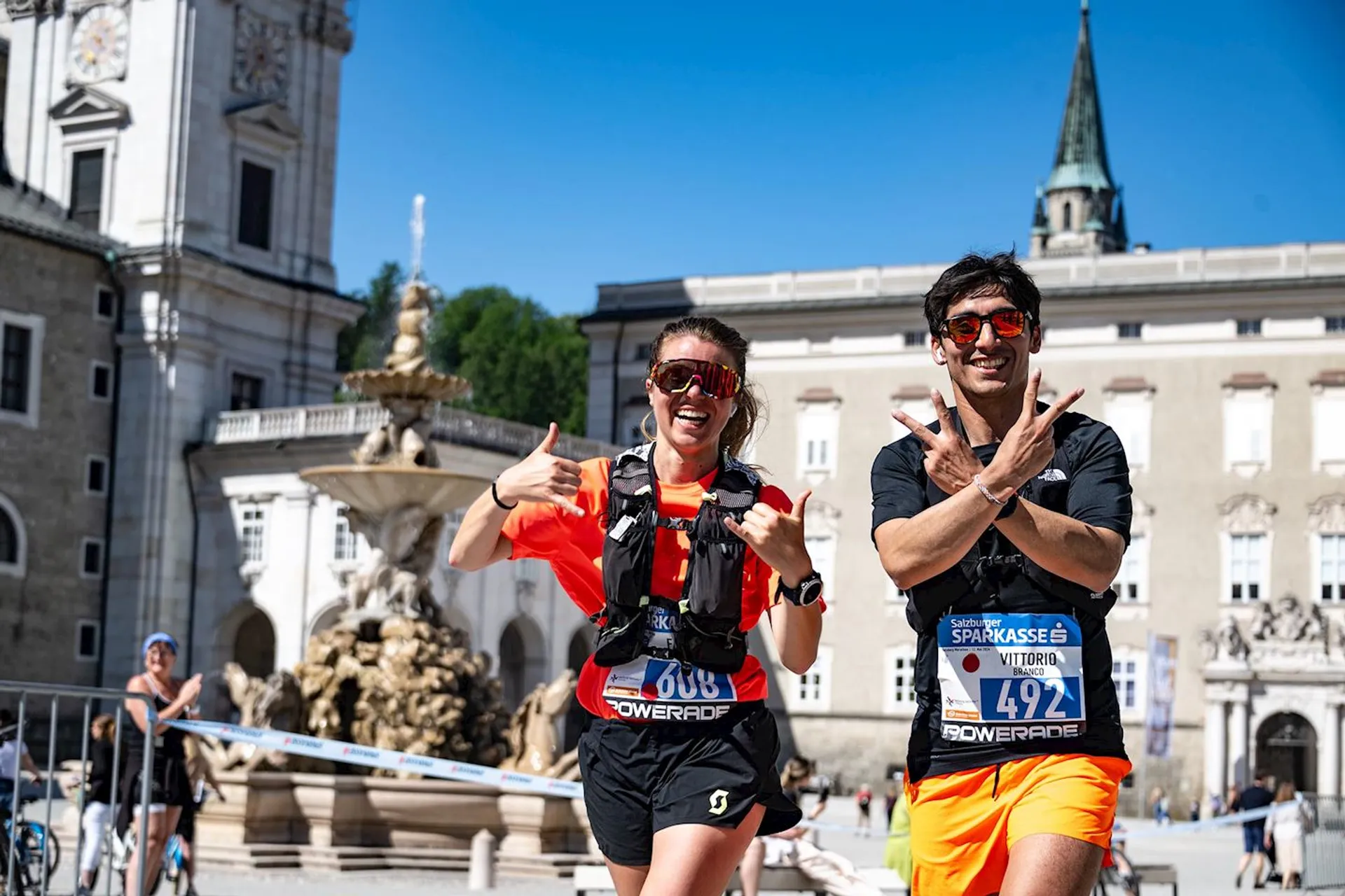 The image shows two people in athletic gear participating in a racing event. They are smiling and gesturing with their hands while wearing race bibs with numbers. The background features a fountain and historic architecture, indicating they might be in a European city. The atmosphere appears to be a fun, outdoor event.