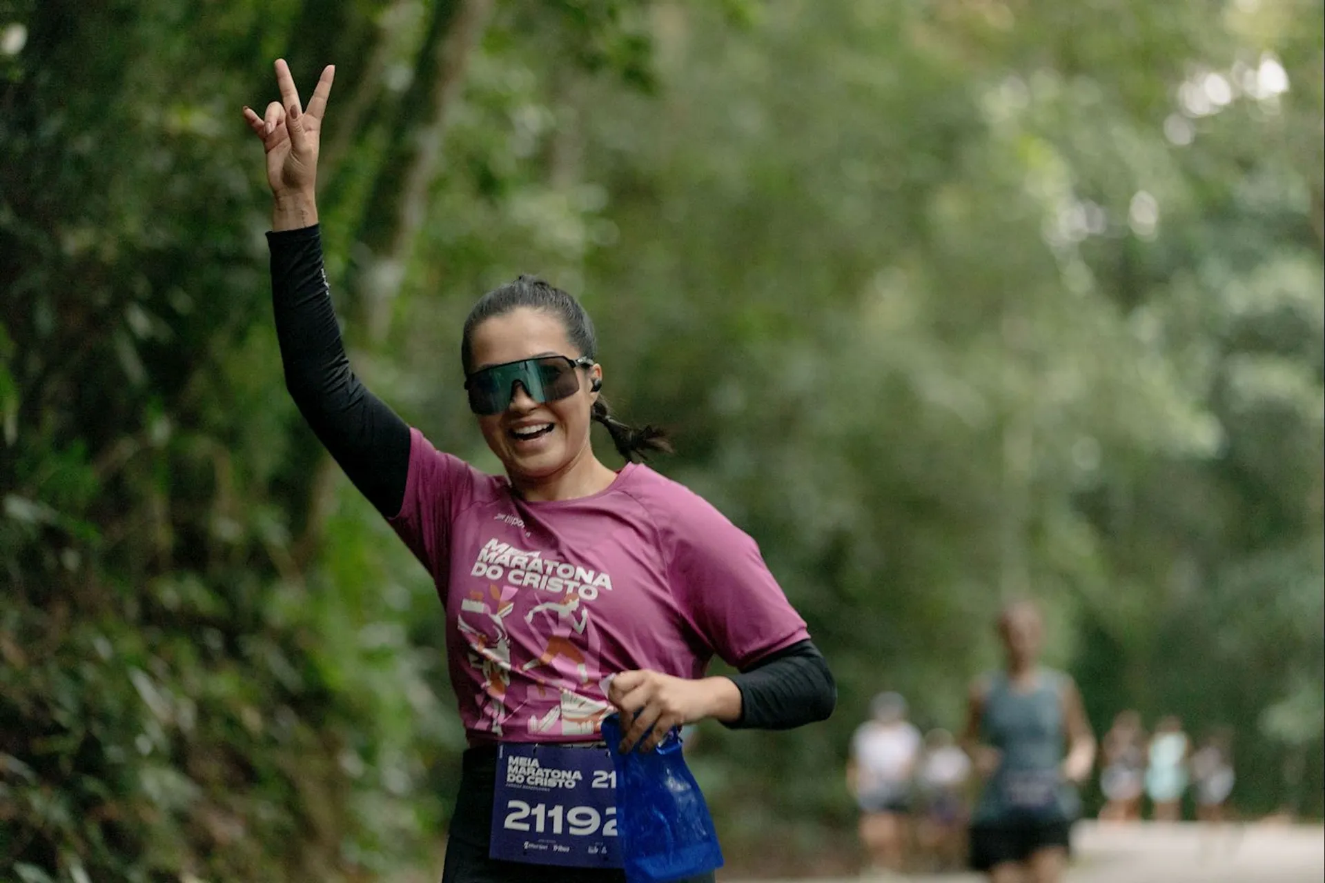The image shows a person wearing a pink running shirt and large sunglasses, holding up a peace sign with one hand, while running in an outdoor setting. There are trees and other runners in the background. The person appears to be participating in a marathon or race, as indicated by the race bib.