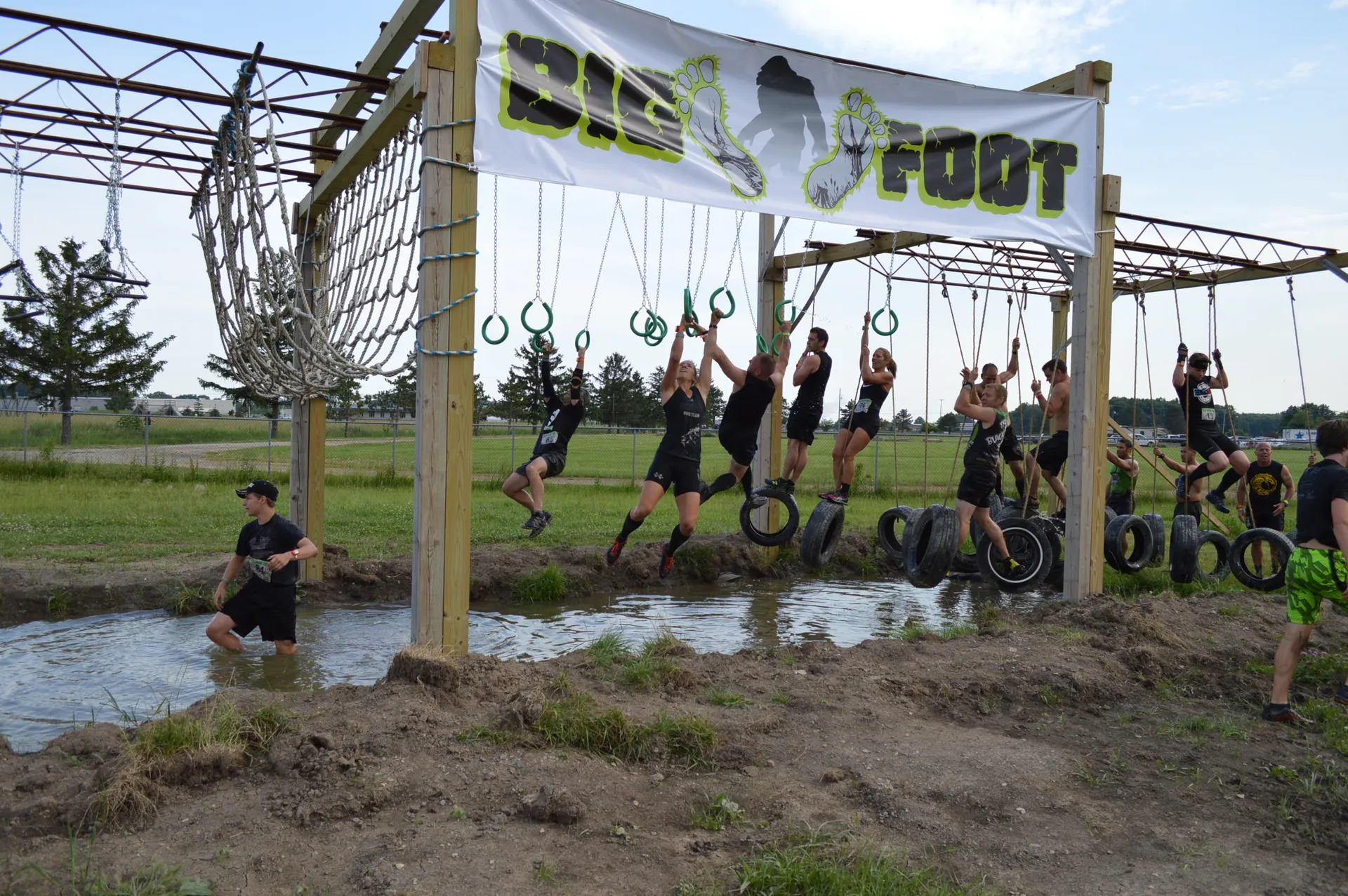 The image shows an outdoor obstacle course with people engaging in a physical activity. A