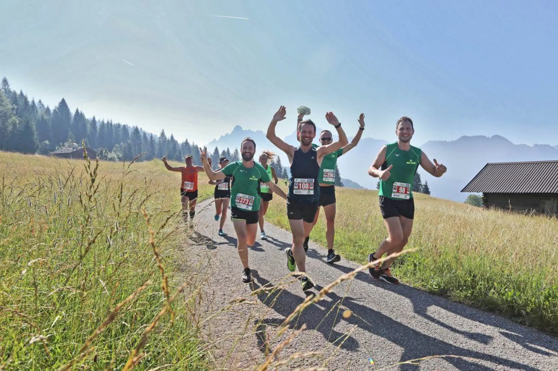 This image features a group of people participating in a running event. They are wearing athletic gear, including running shorts, shirts, and race bibs - which indicates they are part of an organized run or race. The runners appear to be in high spirits, with some raising their hands in a gesture of excitement or victory. They are running on a grassy field, and in the background, there is a picturesque landscape of mountains with a clear sky above, suggesting the event is taking place in a scenic, possibly alpine, environment. Overall, it's a dynamic and cheerful scene with the participants enjoying their athletic endeavor in a beautiful outdoor setting.