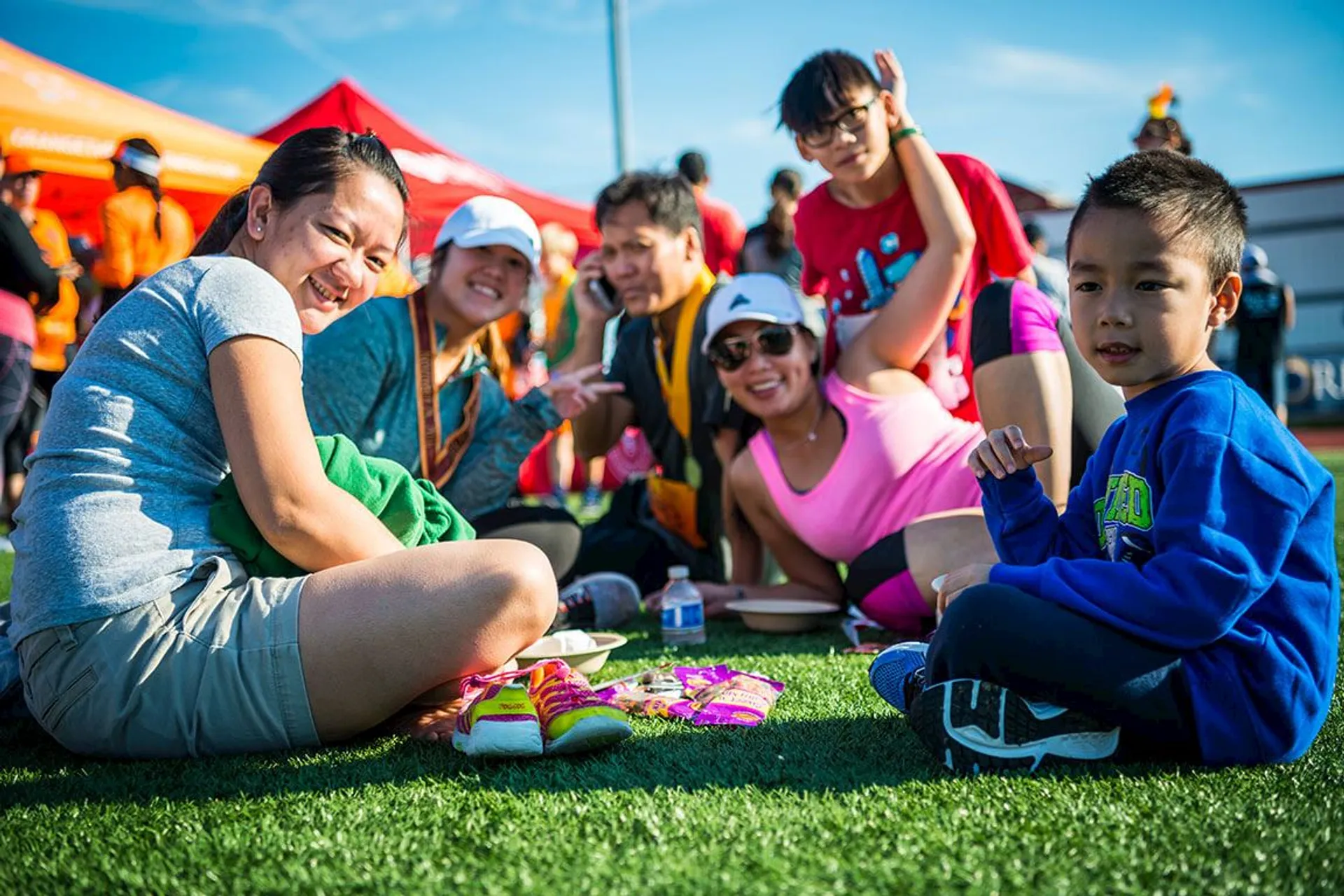 The image shows a group of people sitting on the grass, likely at an outdoor event or gathering. They appear to be enjoying a meal or snacks, with items such as water bottles and food packets visible. Some are wearing casual clothing, including athletic attire. The background features red tents and a clear blue sky. Everyone seems to be in a cheerful mood.