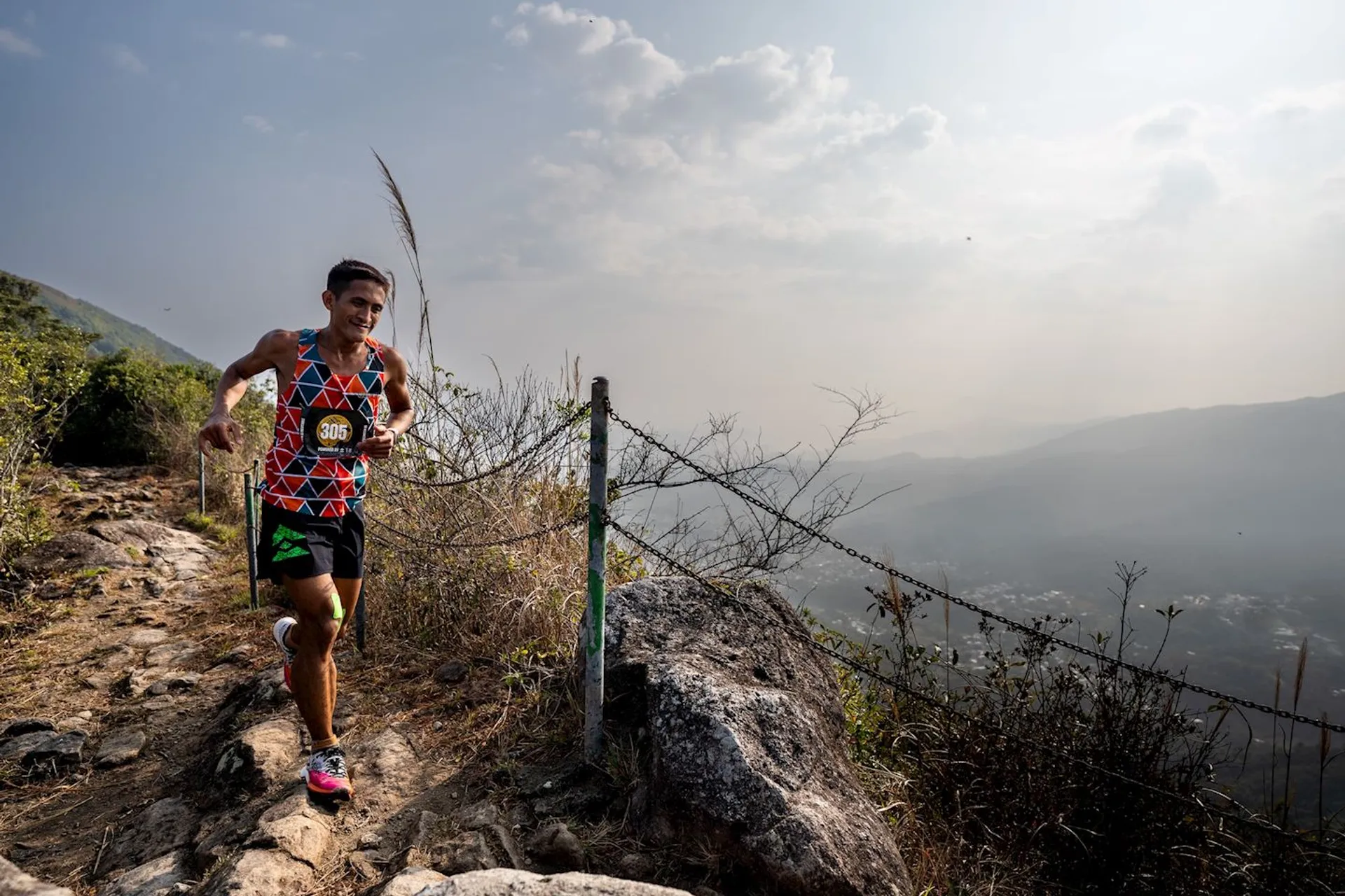 The image shows a person running on a rocky, uphill trail. The runner is wearing a colorful athletic outfit and is next to a fence with a scenic, mountainous background. The sky is partly cloudy, and the scenery gives a sense of elevation and outdoor adventure.