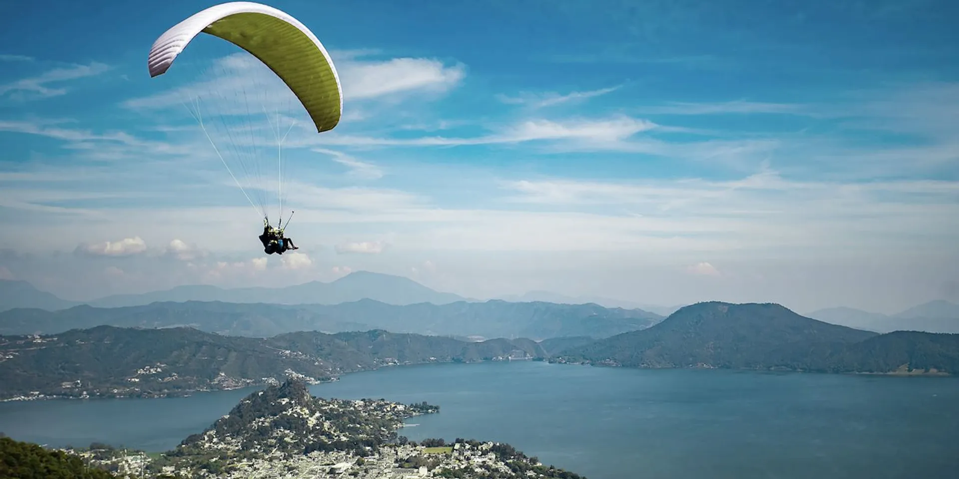 In this image, there is a person paragliding above a scenic landscape. The paraglider is using a canopy that appears to be yellow and white, and they are flying over a body of water and an area with rolling hills or mountains. The skies are clear with very few clouds, making it an ideal day for such an activity. In the foreground, there appears to be a cluster of buildings, possibly a small town, adjacent to the water. The setting is serene and picturesque, suggesting a great location for outdoor sports and recreation.