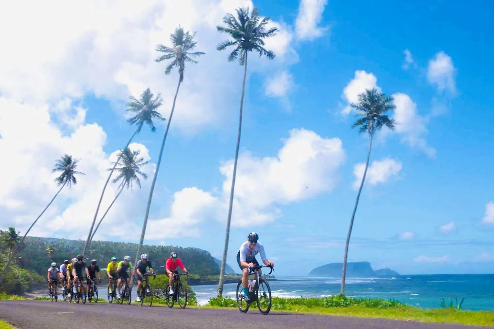 The image shows a group of cyclists riding along a coastal road. Tall palm trees and a clear blue sky with a few clouds are visible. In the background, there's a view of the ocean and a distant landmass. The setting is tropical and scenic.