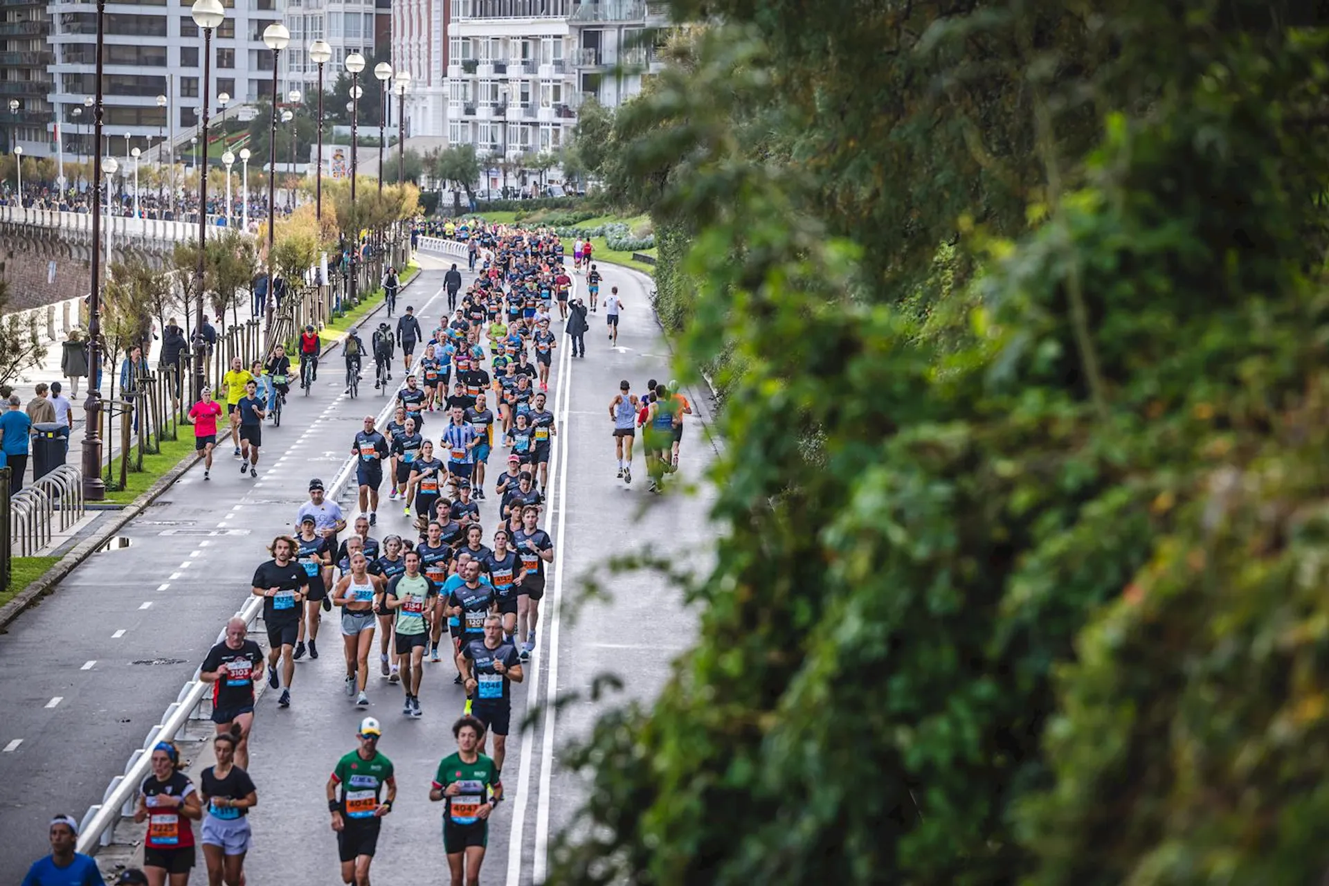 The image shows a large group of people running in a marathon or race event along a city street. They appear to be on a designated path with greenery and high-rise buildings visible in the background. The area is well-organized, likely part of a planned running event.
