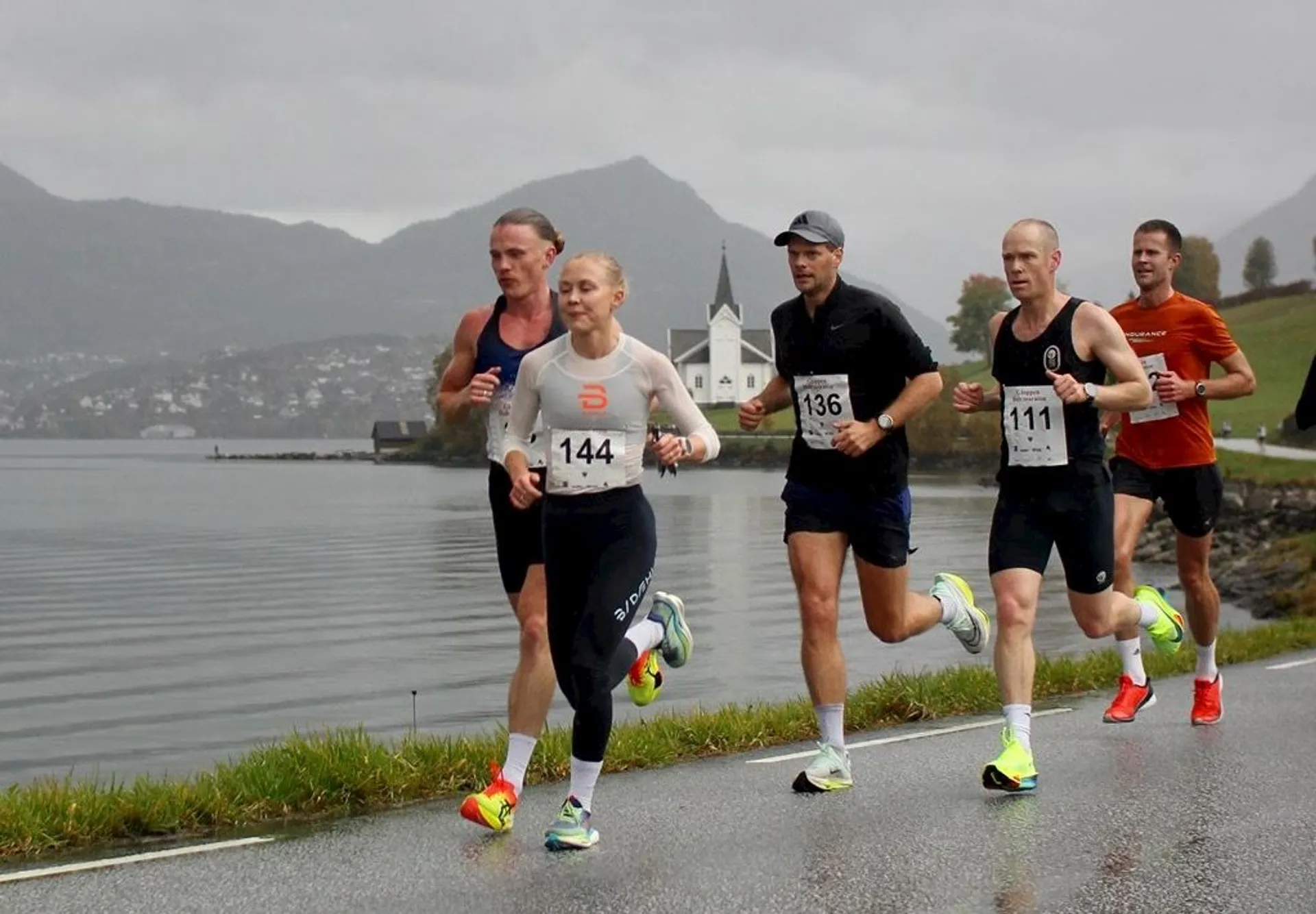 The image shows a group of runners participating in a race along a scenic route. They are running beside a body of water with mountains and a church visible in the background. The runners all have numbered bibs and appear to be focused and determined. The weather looks overcast and the ground appears wet, indicating recent rain.