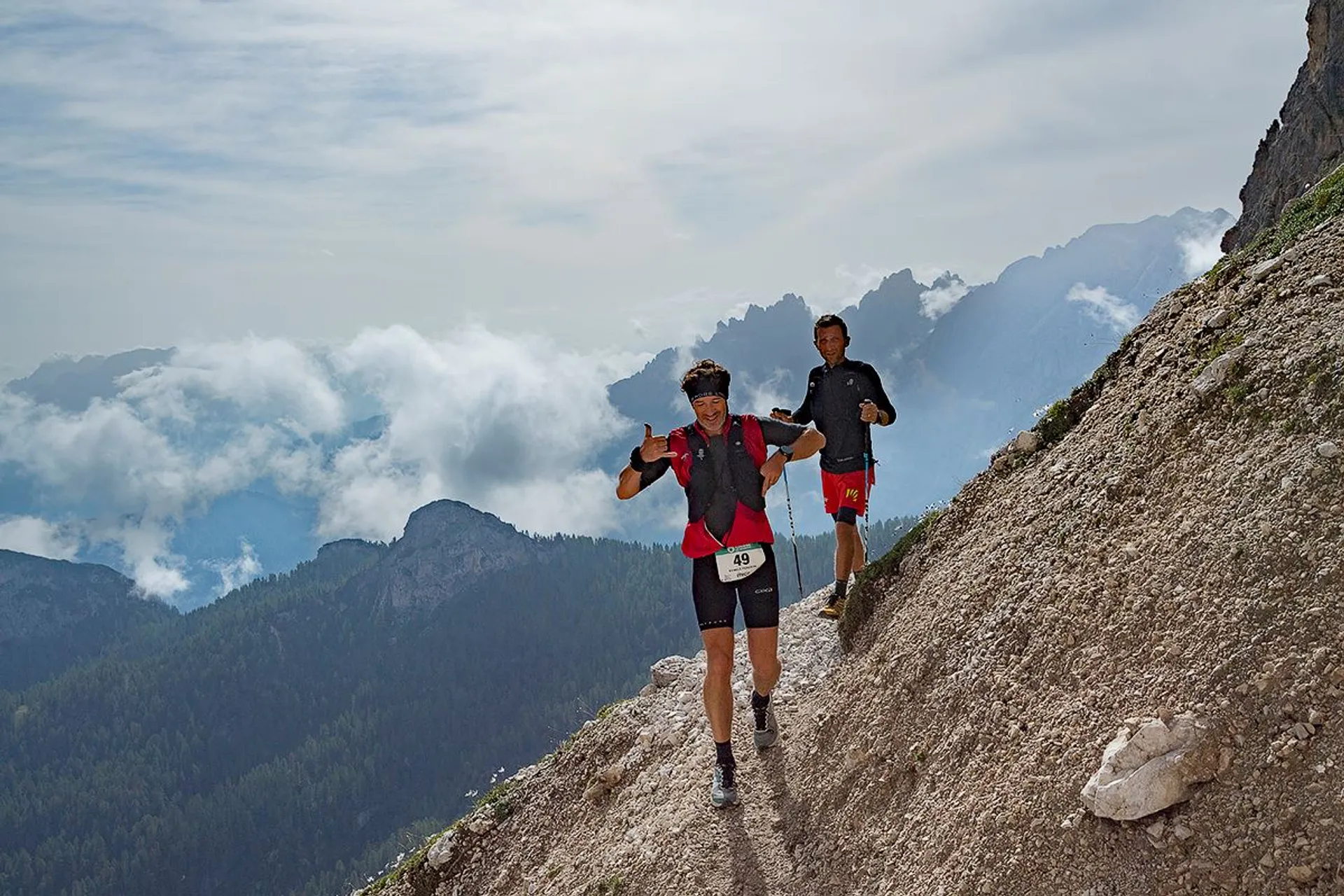 This image shows two people running on a narrow mountain trail. They are dressed in athletic gear and appear to be participating in a trail or mountain race. The background features a scenic view of mountains and clouds.