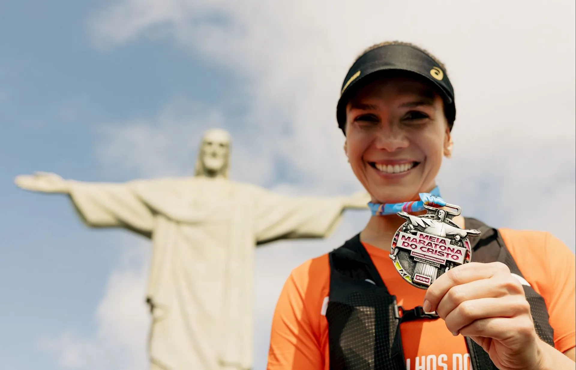 The image shows a person smiling and holding a medal with a statue in the background. The statue resembles the famous Christ the Redeemer in Rio de Janeiro. The person appears to be celebrating a race or event, possibly related to the marathon indicated on the medal.