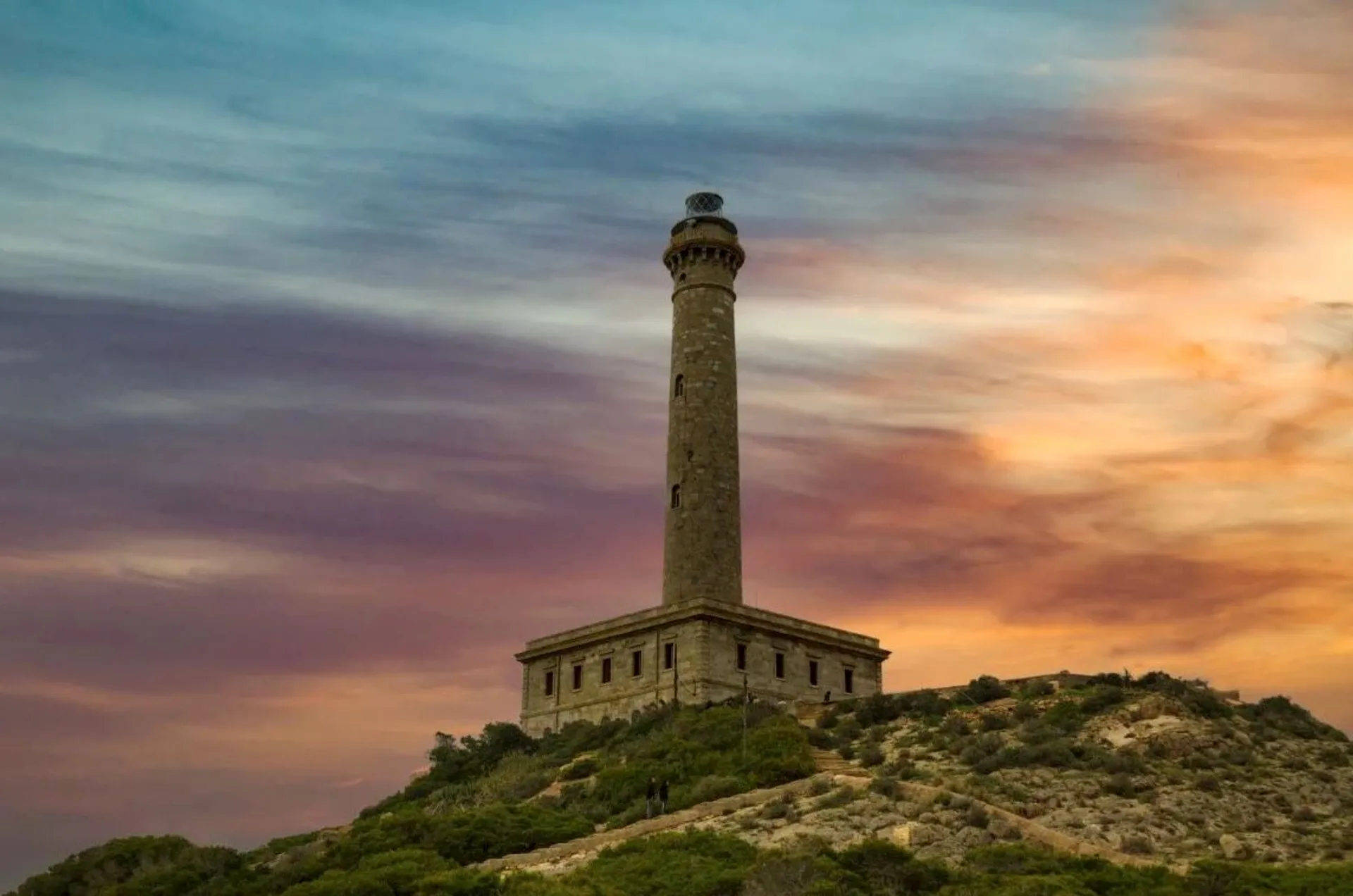 The image shows a lighthouse situated on a rocky hill with a colorful sky at sunset or sunrise. The building has a tall, cylindrical tower with a light at the top, and there's surrounding vegetation at the base of the hill.