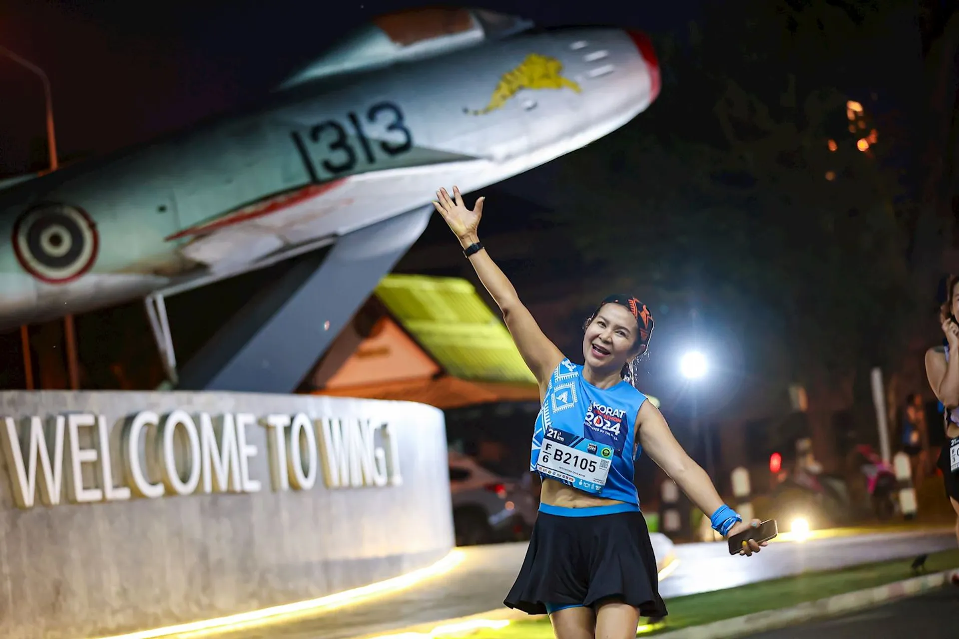 The image shows a person posing happily during what appears to be a running event, based on their attire and race number bib. In the background, there's a fighter jet displayed on a platform with the text "WELCOME TO WING 1." The scene is likely set at an airbase or aviation-themed location.