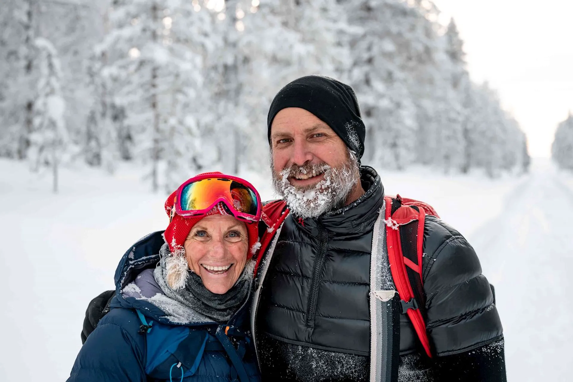 This image shows two people outdoors in a snowy environment. They are both dressed in winter clothing, including hats. One person is wearing goggles and has snow on their face, and the other has snow on their beard. They appear to be enjoying a winter activity, possibly skiing or snowshoeing, given the attire and surroundings. The background features snow-covered trees.