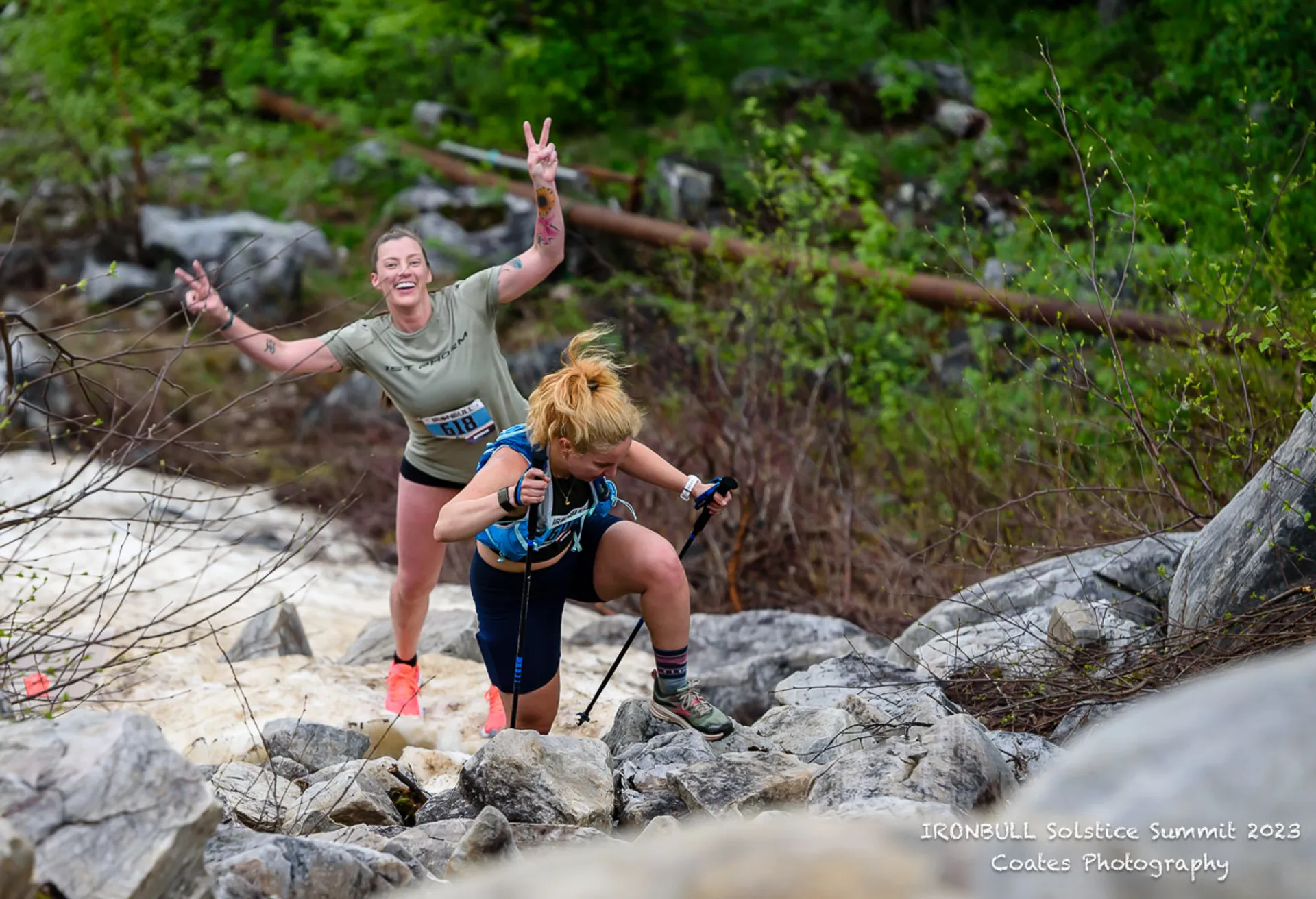 The image shows two people participating in an outdoor trail or adventure race. They're climbing over rocky terrain, with one person using trekking poles and the other making a peace sign gesture. They're dressed in athletic gear and appear to be enjoying the activity. The setting is natural and rugged, possibly part of a mountain or wooded area.