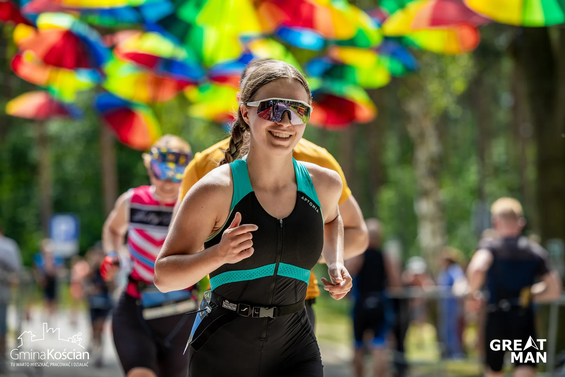 The image shows people participating in a running event. They are wearing athletic gear and sunglasses. There are colorful umbrellas hanging above them, creating a festive atmosphere. The setting appears to be outdoors, possibly in a park or wooded area.