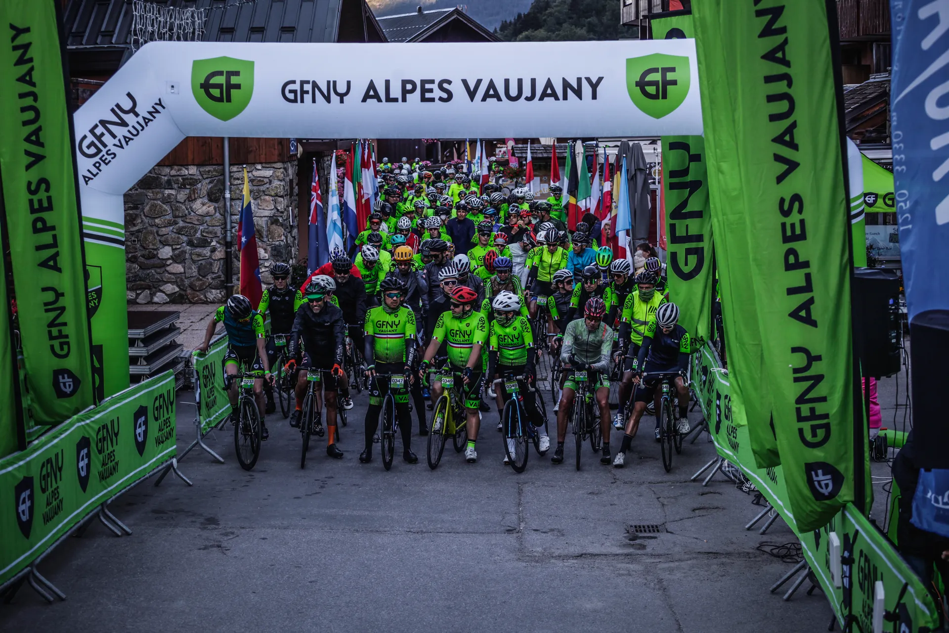 The image shows a group of cyclists at the starting line of an event called GFNY Alpes Vaujany. The cyclists are wearing green jerseys, and there are banners and flags from various countries displayed. The setting appears to be an organized race or cycling event in a mountainous region.