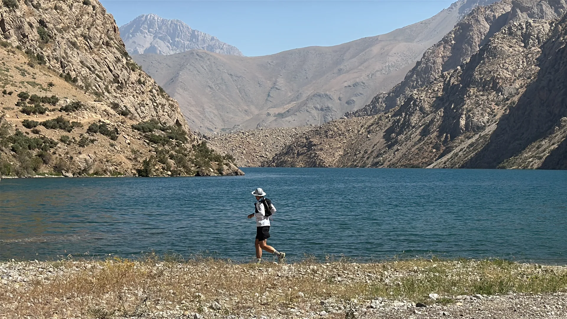 This image shows a person jogging by the shore of a large blue lake surrounded by mountainous terrain. The person is wearing a white shirt, black shorts, and a hat, and is carrying a backpack. The landscape features rocky hills and clear skies.