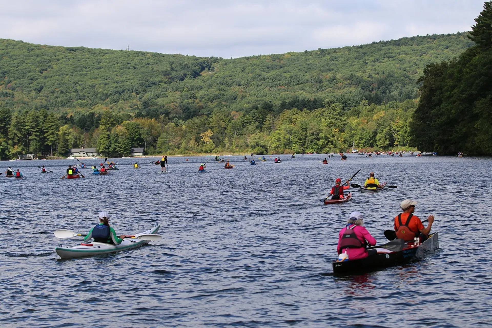 The image shows a body of water with a hilly, forested landscape in the background. In the water, there is a variety of small boats, including kayaks and canoes, with people paddling. The paddlers are wearing life vests, suggesting a focus on safety. The scene seems to be a recreational day out on the water, where people are enjoying the outdoor activities and the natural environment.