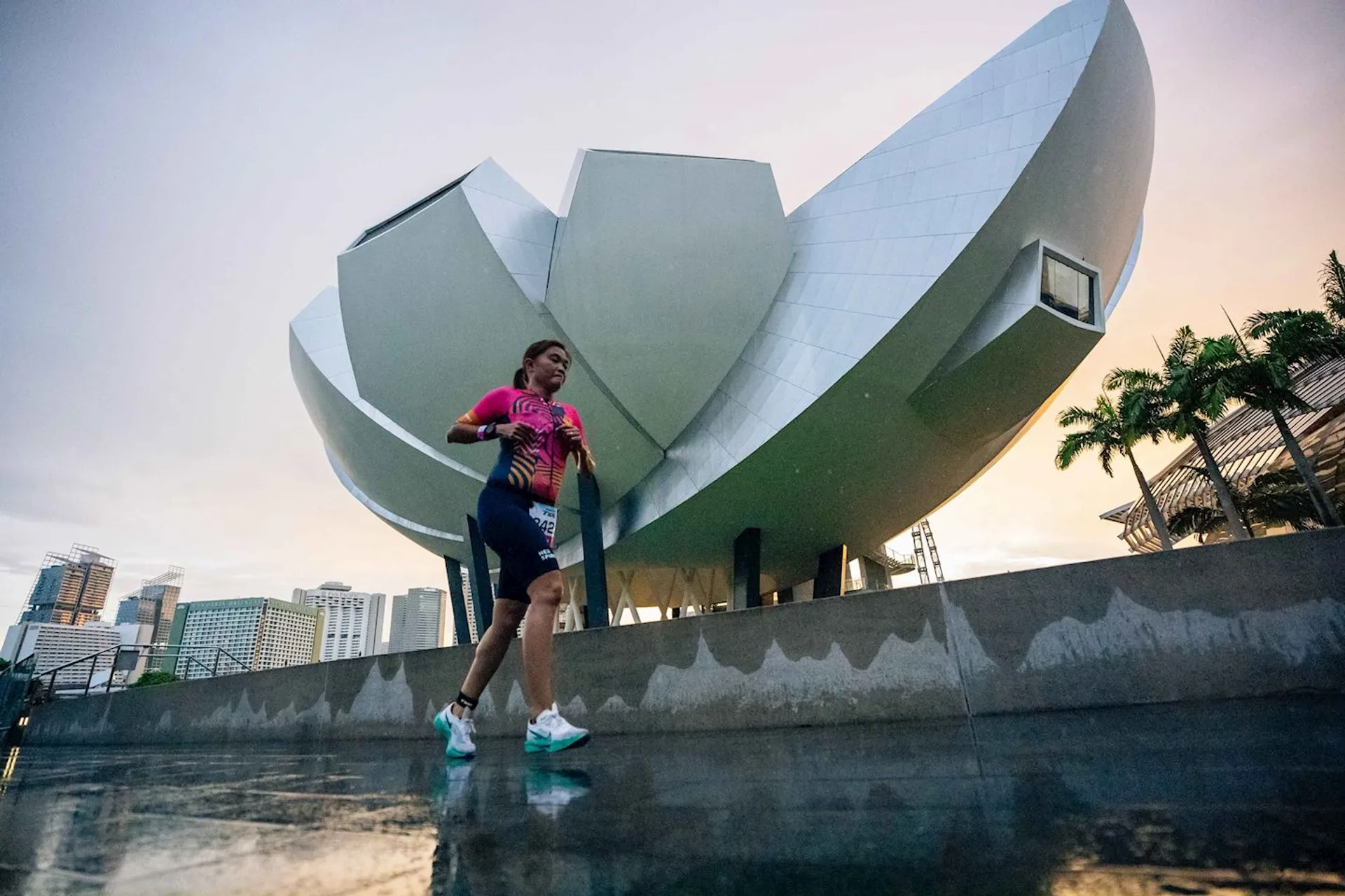 The image shows a person walking in front of a distinctive modern building with an architectural design resembling an open lotus or flower. It appears to be during a run or exercise, as the person is dressed in athletic wear. The background includes a cityscape with several tall buildings and palm trees, suggesting an urban setting.
