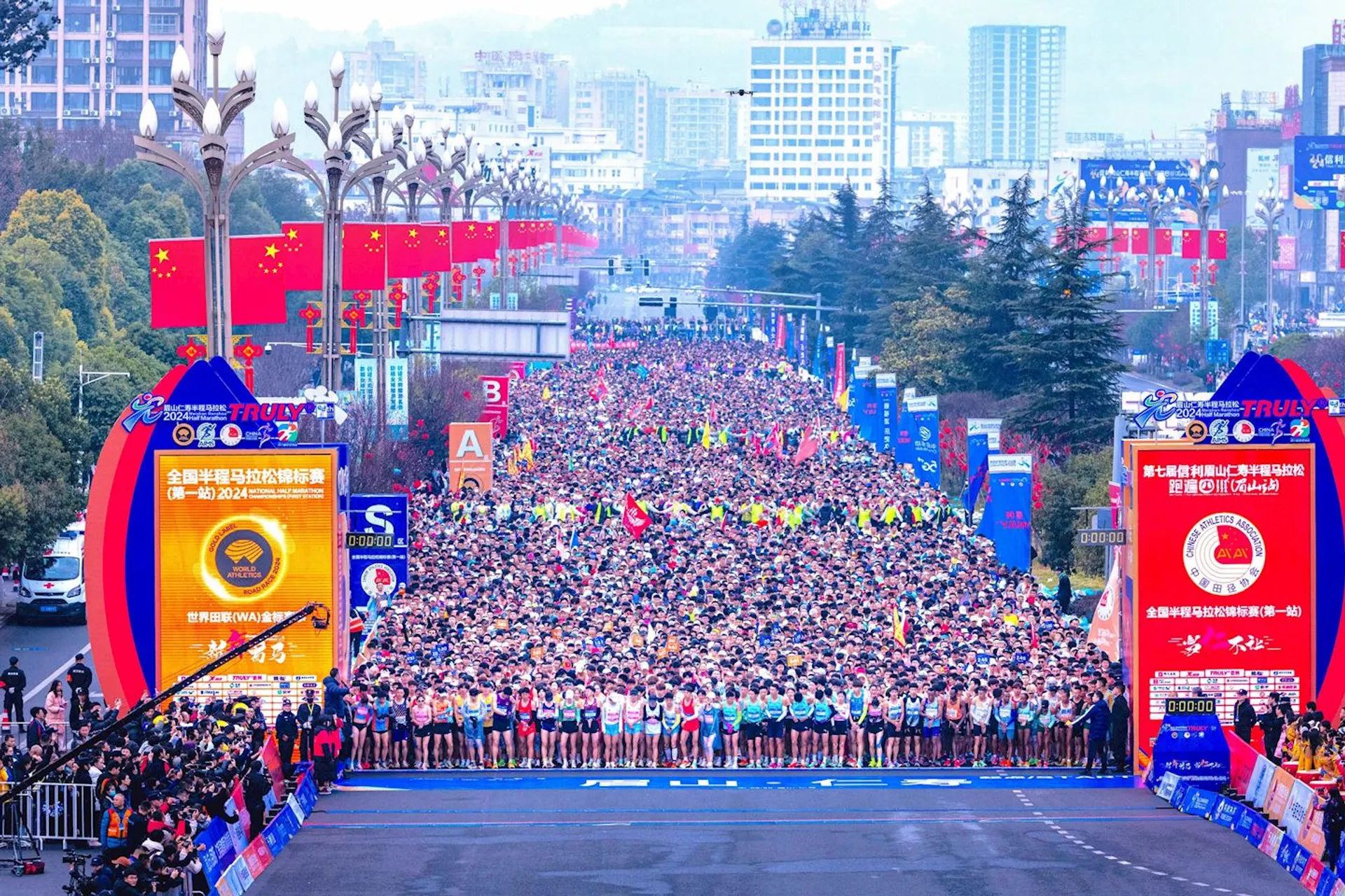 The image shows a large crowd of marathon runners lined up at the starting line of a race. There are numerous participants in sportswear, and the scene includes organizers and spectators along the sides. There are red banners and displays with Chinese characters, indicating it's a marathon event in China. The cityscape in the background features tall buildings and trees.