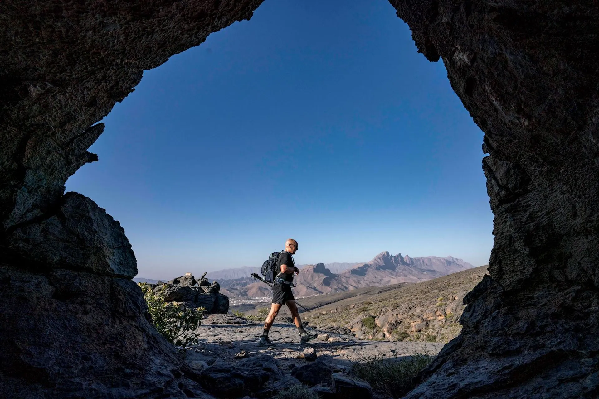 The image depicts a person hiking through a rocky landscape, framed by the opening of a cave or rocky arch. In the background, there are mountains under a clear blue sky. The person is wearing a backpack and hiking gear.