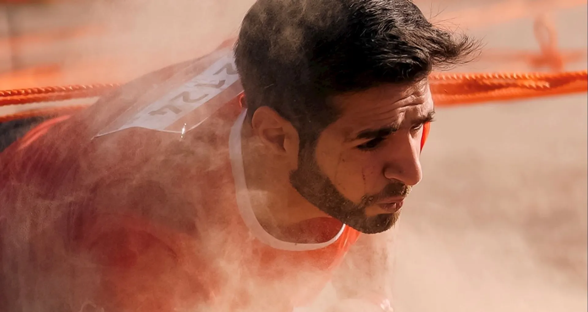 The image shows a person participating in an obstacle race. They are crawling under an obstacle with orange ropes, and there is dust around them, creating an intense and dynamic atmosphere. The person is wearing a bib number on their back.