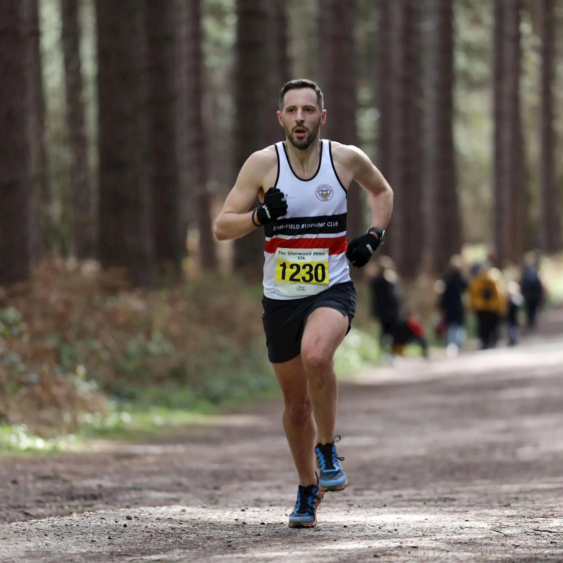 The image shows a man running an outdoor race on a path through a wooded area. He's wearing a white sleeveless running shirt with logos, black gloves, black running shorts, and blue running shoes. He has a runner's bib attached to his shirt with the number 1230. His expression is focused, and he appears to be exerting effort. There are other runners and spectators in the background. The environment suggests it might be a marathon or trail running event.