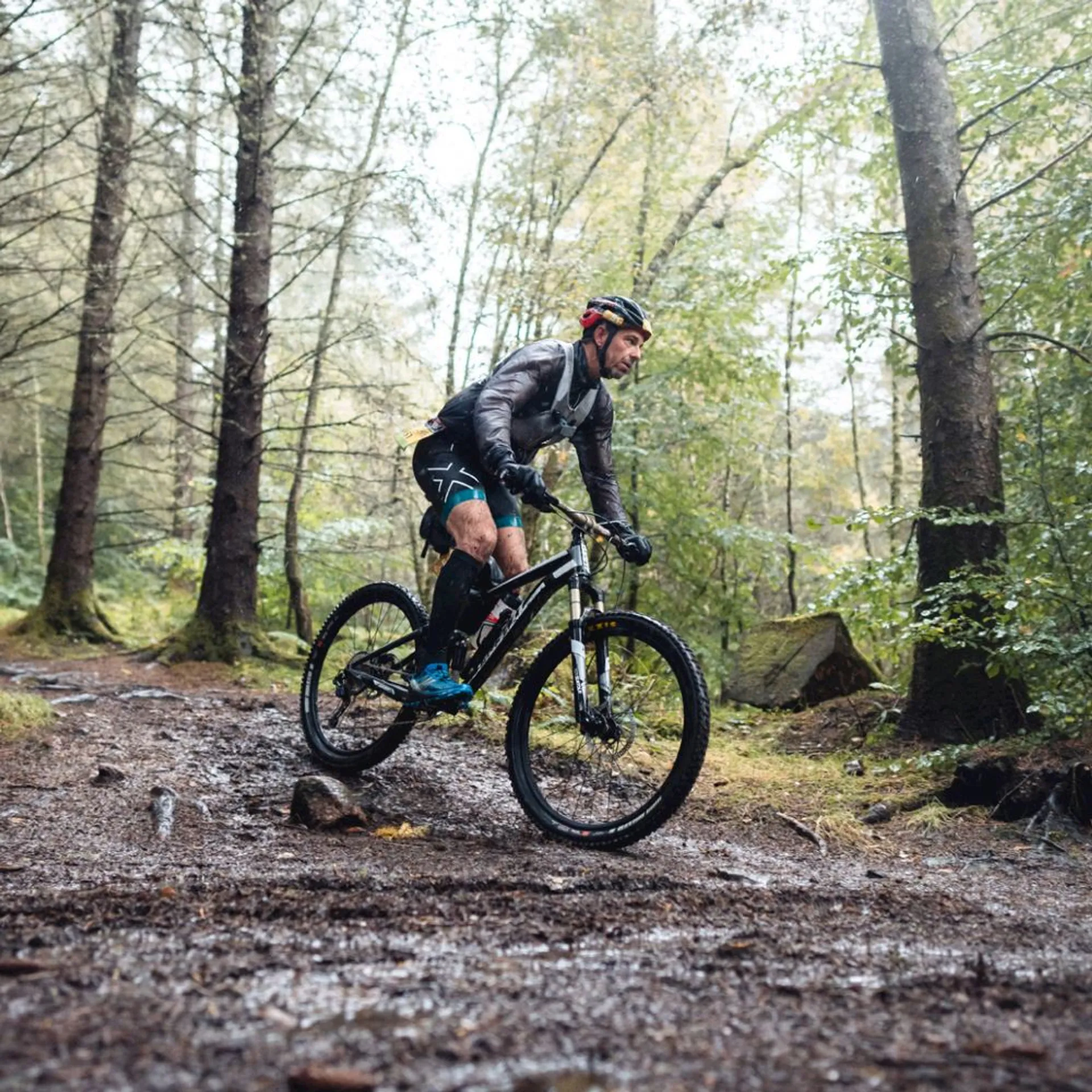 The image shows a person riding a mountain bike through a forested area. The ground appears to be wet, possibly due to recent rain, as evidenced by the moist soil and puddles on the trail. The biker is dressed in sporty attire suitable for cycling, which includes a helmet for safety, gloves, a jacket, and specialized shoes. The dense trees and the natural scenery suggest that this is a trail well-used for mountain biking or outdoor activities. The biker seems to be in motion as they navigate through the rough trail filled with rocks and fallen leaves.