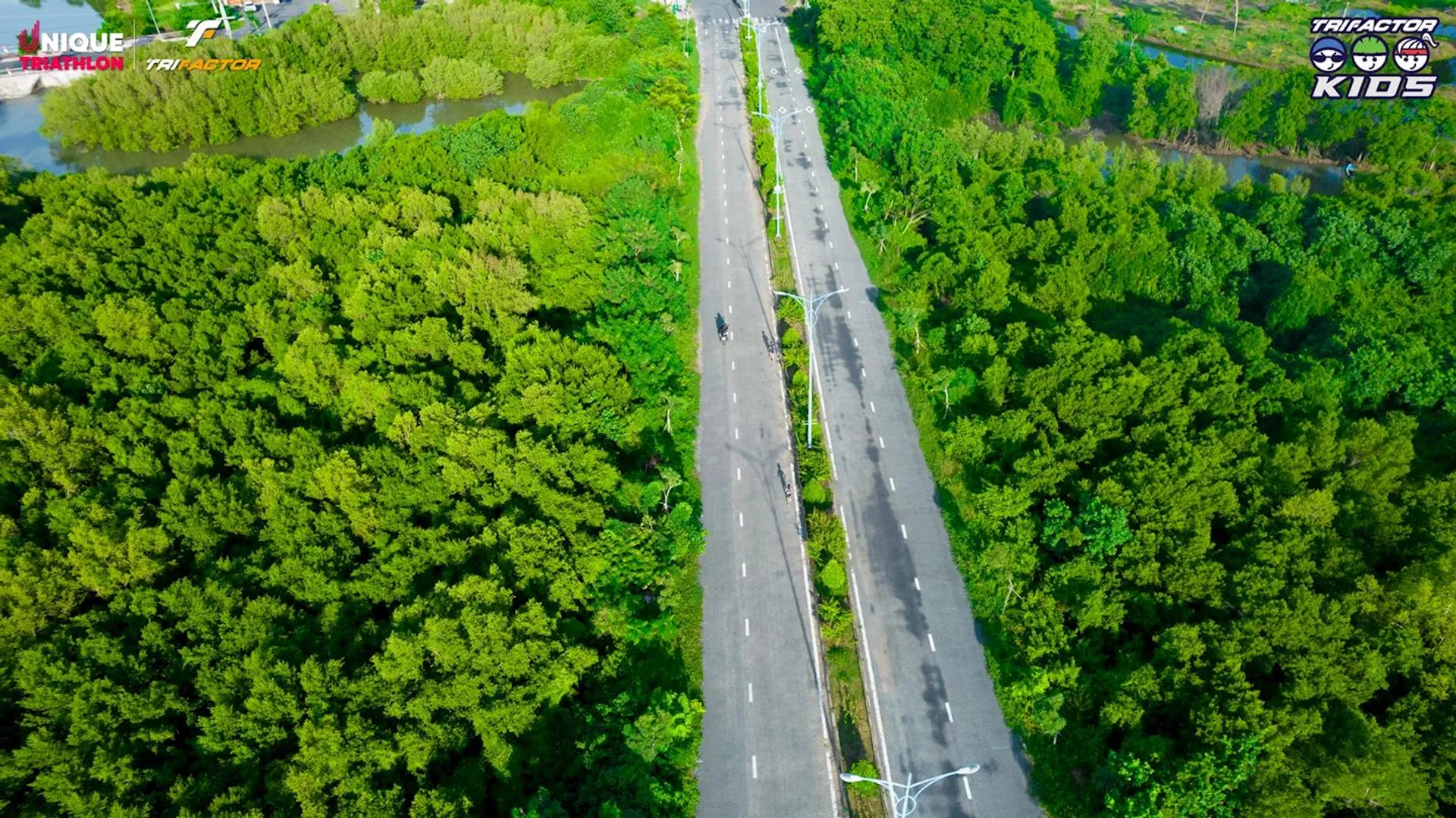 This image shows a top-down view of a road surrounded by lush, green vegetation. The road appears to be mostly empty except for some cyclists riding along it. There are also logos related to a triathlon event visible in the corners of the image.