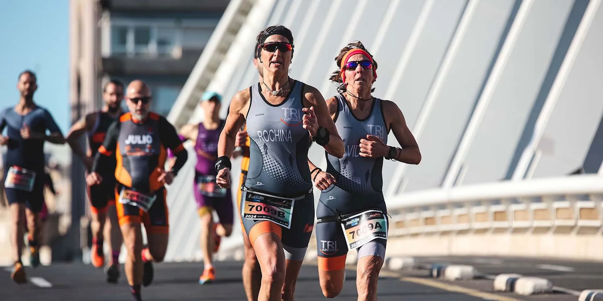The image shows a group of athletes participating in a running event, possibly a triathlon, given their attire which includes racing suits with sponsor logos and race numbers visible. The two athletes in the foreground are wearing sunglasses and race belts with their numbers attached, suggesting this could be the running segment of a triathlon. The location appears to be a bridge or a similar structure, considering the curved architecture in the background, and the weather is sunny and clear. The competitors are focused and appear to be running at a steady pace.