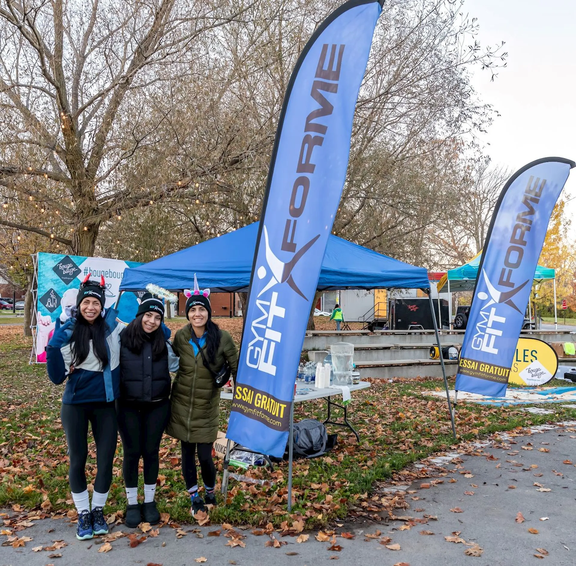 The image shows three people standing outdoors near a small setup, which includes two blue banners with "FIT FORME" written on them. There are tents and tables behind them, and the ground is covered in fallen leaves, indicating it might be autumn. The group is posing for the photo, and they appear to be participating in or attending an event.