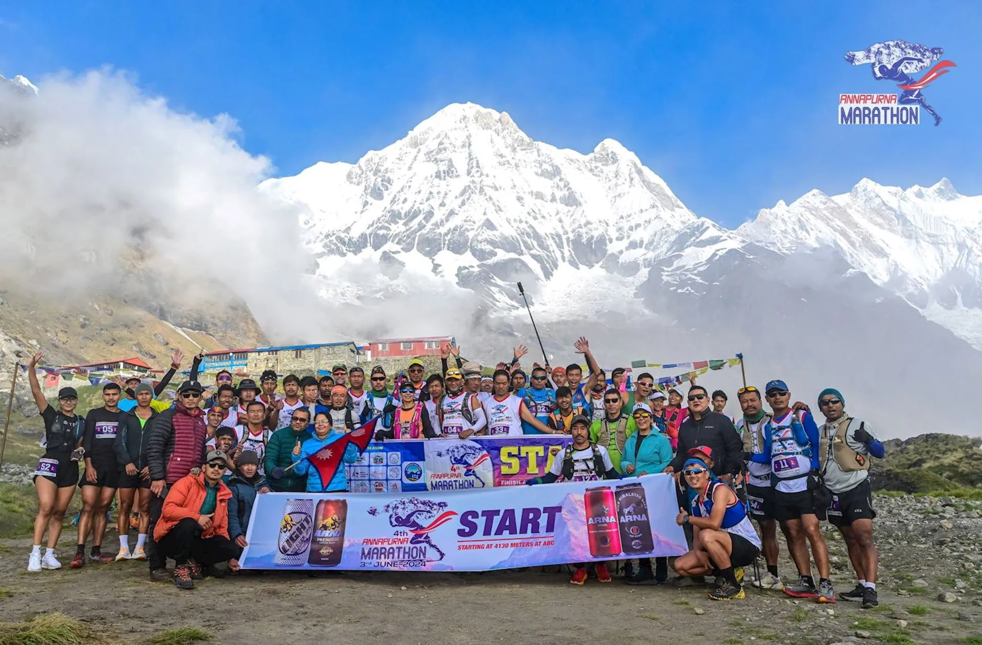 This image shows a large group of people gathered in front of a snowy mountain peak. They are posing with banners related to the "Annapurna Marathon" and some are holding flags. The scene is outdoors with mountainous terrain, likely at a high altitude. The group appears to be celebrating or preparing for a running event.