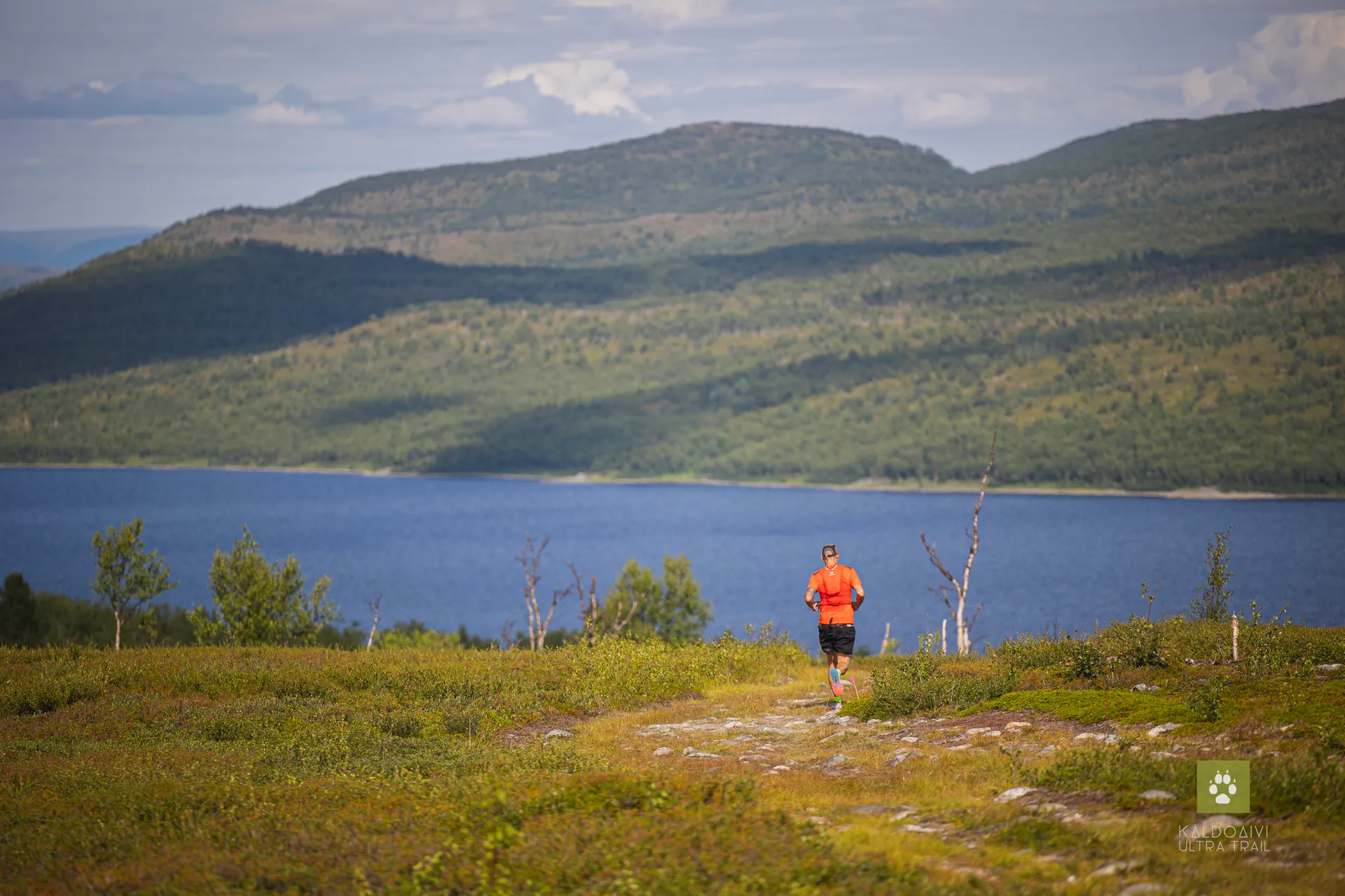 This image shows a person running on a trail in a natural landscape. The runner is wearing a bright orange top and black shorts, which stand out against the natural colors of the scene. The runner is in focus while the background is slightly blurred, highlighting their movement through the landscape. The scenery includes rolling hills or low mountains, and there is a large body of water, such as a lake or wide river, in the distance. Sparse vegetation and a few small trees suggest this might be a high-altitude or subalpine environment. The weather appears to be clear with a mix of clouds, suggesting it's a nice day for outdoor activities.