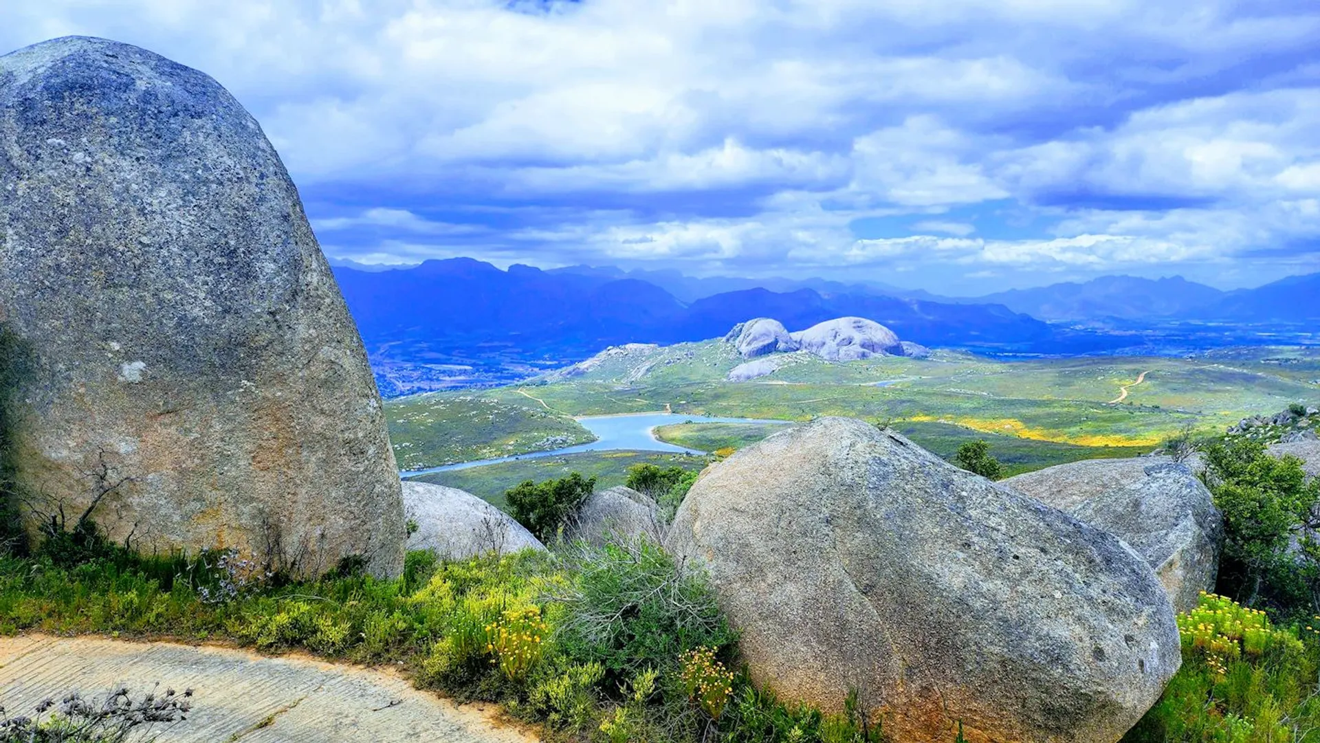The image features a scenic landscape with large rounded boulders in the foreground, overlooking a valley. A body of water can be seen in the middle ground, snaking through the valley with patches of greenery and fields around it. In the distance, there are mountain ranges under a partly cloudy blue sky. The overall setting looks peaceful and natural, with no signs of urban development visible in this view.
