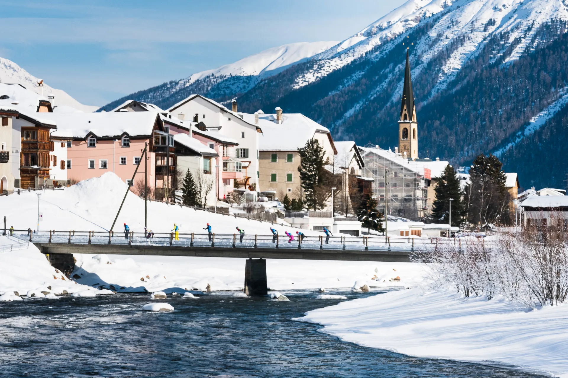 The image depicts a snowy winter scene in a small town or village. There's a bridge crossing over a river with several people walking across it. In the background, there are snow-covered buildings and a tall church steeple. Mountains covered in snow rise up behind the town, creating a picturesque alpine setting.