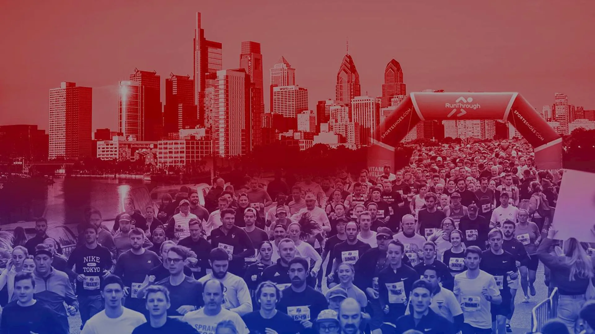 The image shows a large group of people participating in a running event. They are running under an inflatable arch with the word "Runthrough" on it. In the background, there is a city skyline with tall buildings. The image has a red and blue overlay effect.