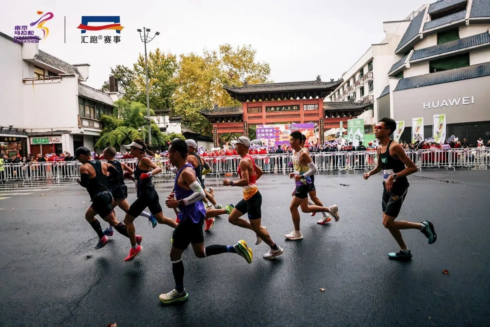 The image shows a group of runners participating in a race. They are running on a wet road, likely during a marathon or similar event. Spectators are behind barriers, and there are buildings in the background. The logos and branding indicate sponsorship and event information.