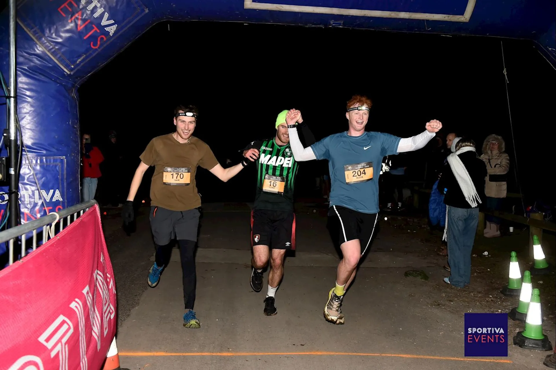 The image shows three runners crossing a finish line together at a nighttime race event. They appear to be celebrating as they hold hands. The event banner has the name "Sportiva Events." A few people are in the background, possibly spectators or event staff.