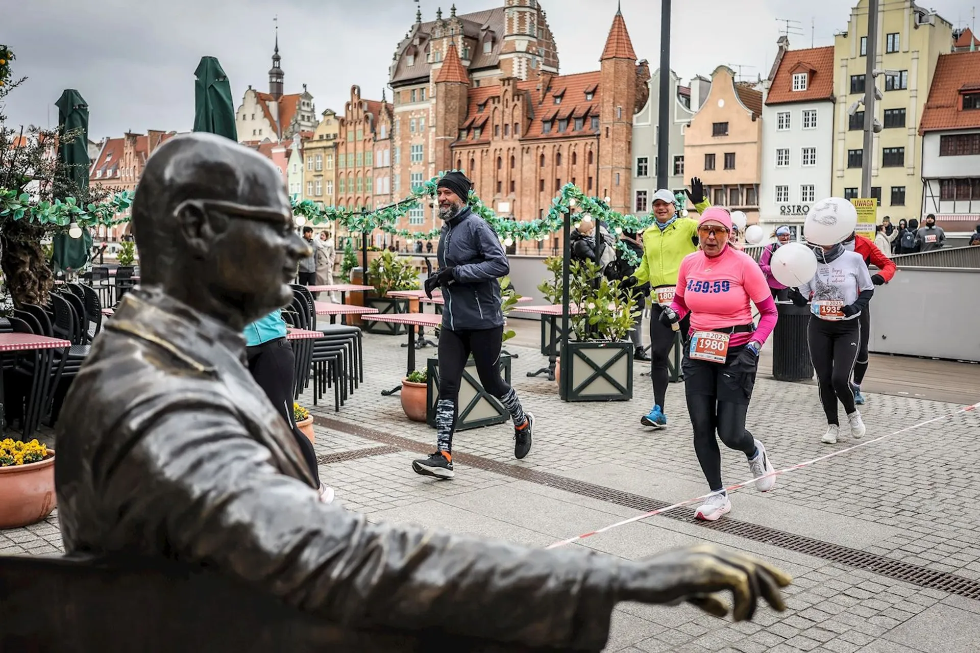 This image shows a group of runners participating in a race, passing by a statue of a seated person. The background features historic buildings, and the scene appears to be in an urban area, possibly during a marathon or organized running event.