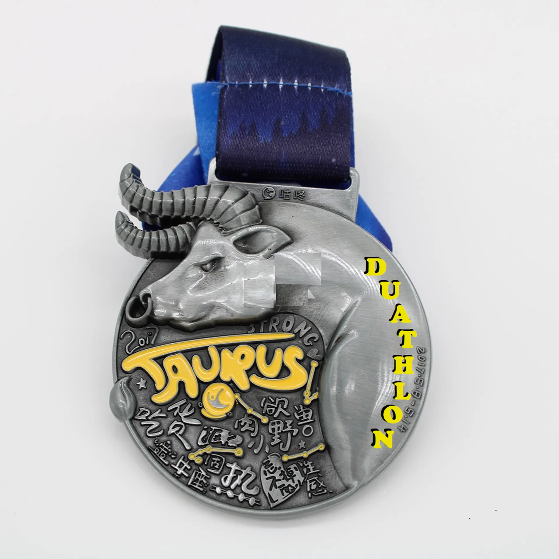 The image shows a medal with a distinctive design. The medal is predominantly silver-colored with some gold and black accents. In the center, there's a three-dimensional relief of a bull's head, which represents the astrological sign Taurus. Above the bull's head, the text "TAURUS" is written in bold, and below the head, the word "DUATHLON" is prominently displayed.

The background of the medal features various symbols or characters that appear to be inspired by East Asian calligraphy, suggesting a cultural or thematic influence in the design of the medal. A blue ribbon is attached to the top of the medal for wearing around the neck, and this ribbon has a darker blue pattern that looks like stylized mountains or waves
