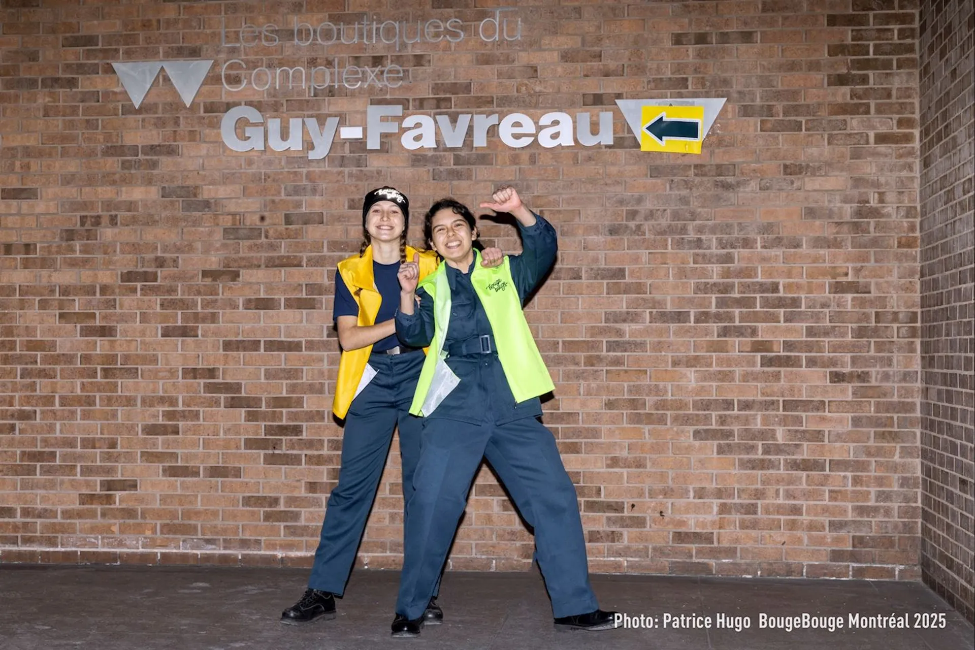 The image shows two individuals posing happily in front of a brick wall that has "Les boutiques du Complexe Guy-Favreau" written on it, along with a directional arrow. They are wearing uniforms with brightly colored vests.