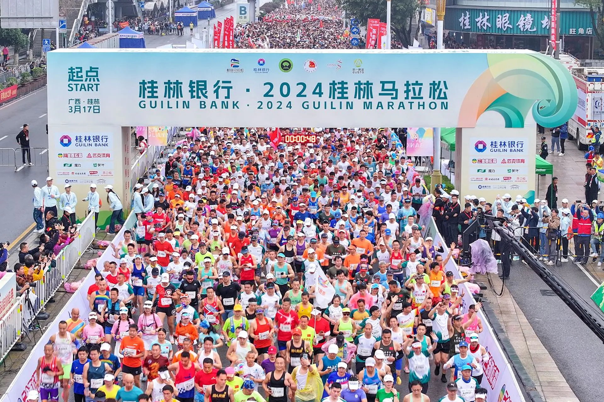 The image shows a large group of runners at the starting line of the 2024 Guilin Marathon. There are banners and signs with text about the event. The scene is vibrant with many participants wearing colorful running gear.