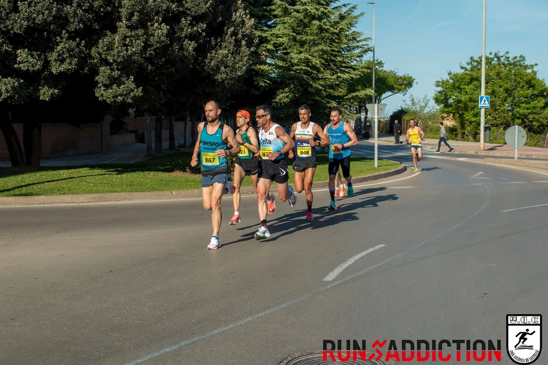The image shows a group of runners competing in an outdoor road race. They are on a street with trees and grass visible in the background. The runners are wearing athletic gear and race numbers. There's branding for "Run Addiction" at the bottom.