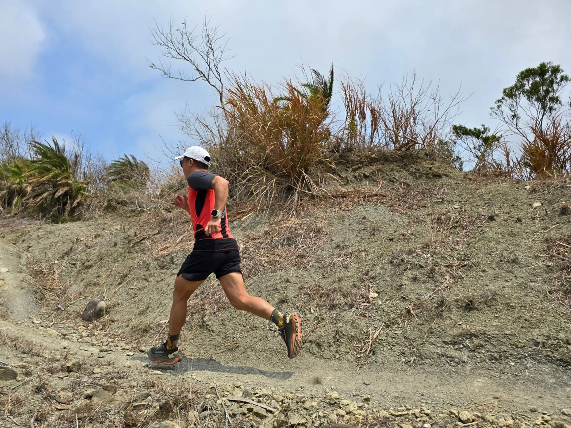 The image shows a person running on a rocky trail. The individual is wearing a red and black outfit with a white cap and has a watch on their wrist. The background includes some dry vegetation and a clear sky.