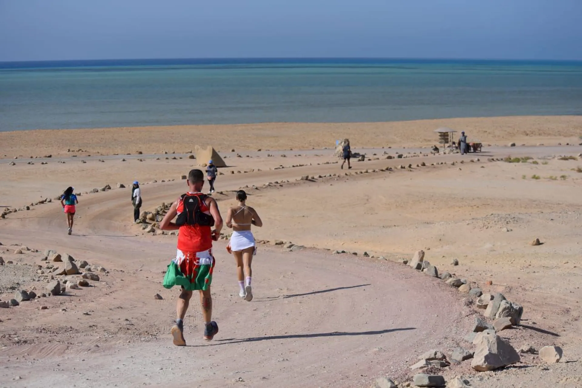 The image shows a group of people running along a path in a desert-like landscape. The runners are heading towards a body of water, possibly a sea or ocean, visible in the background. The landscape is arid with sand and rocks lining the path.